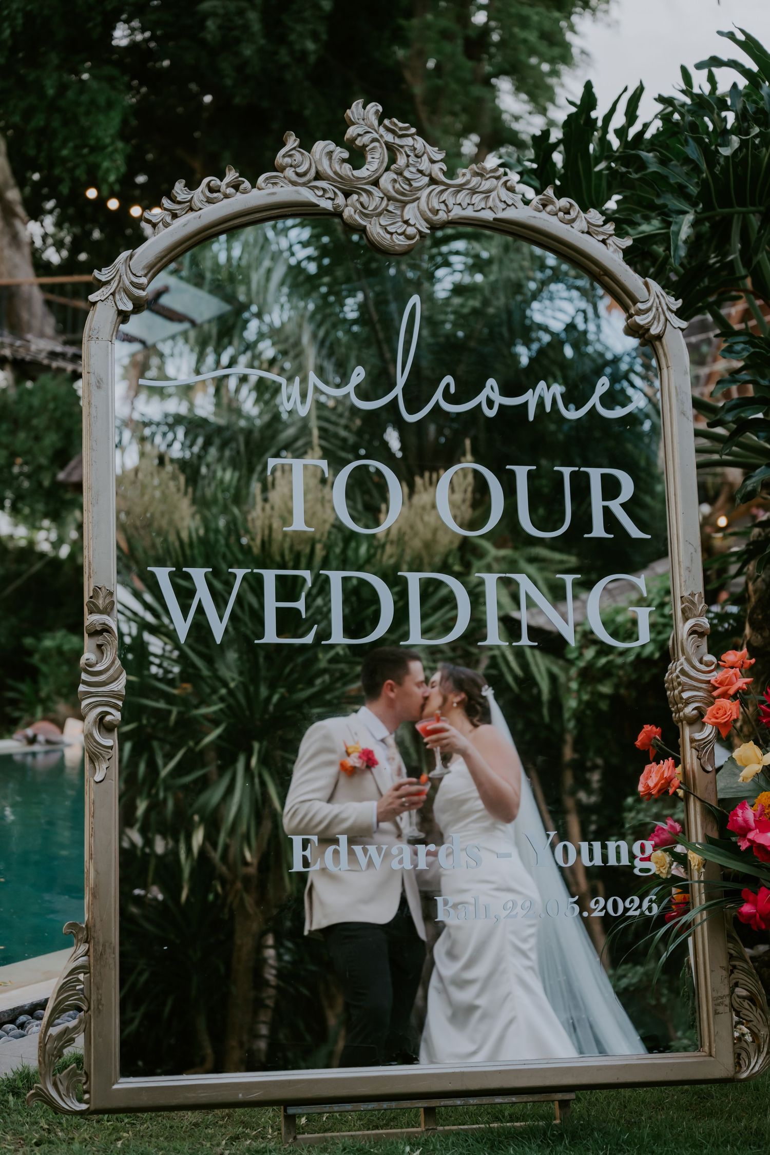Elegant wedding welcome mirror sign with ornate gold frame displaying couple kissing at outdoor garden ceremony.