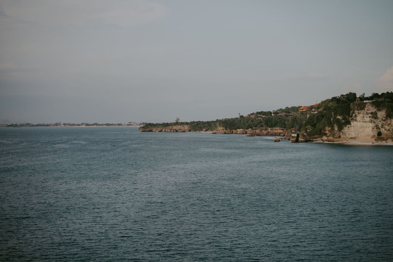 Scenic coastal view with calm blue waters and rocky cliffs lined with lush green trees under a hazy sky.