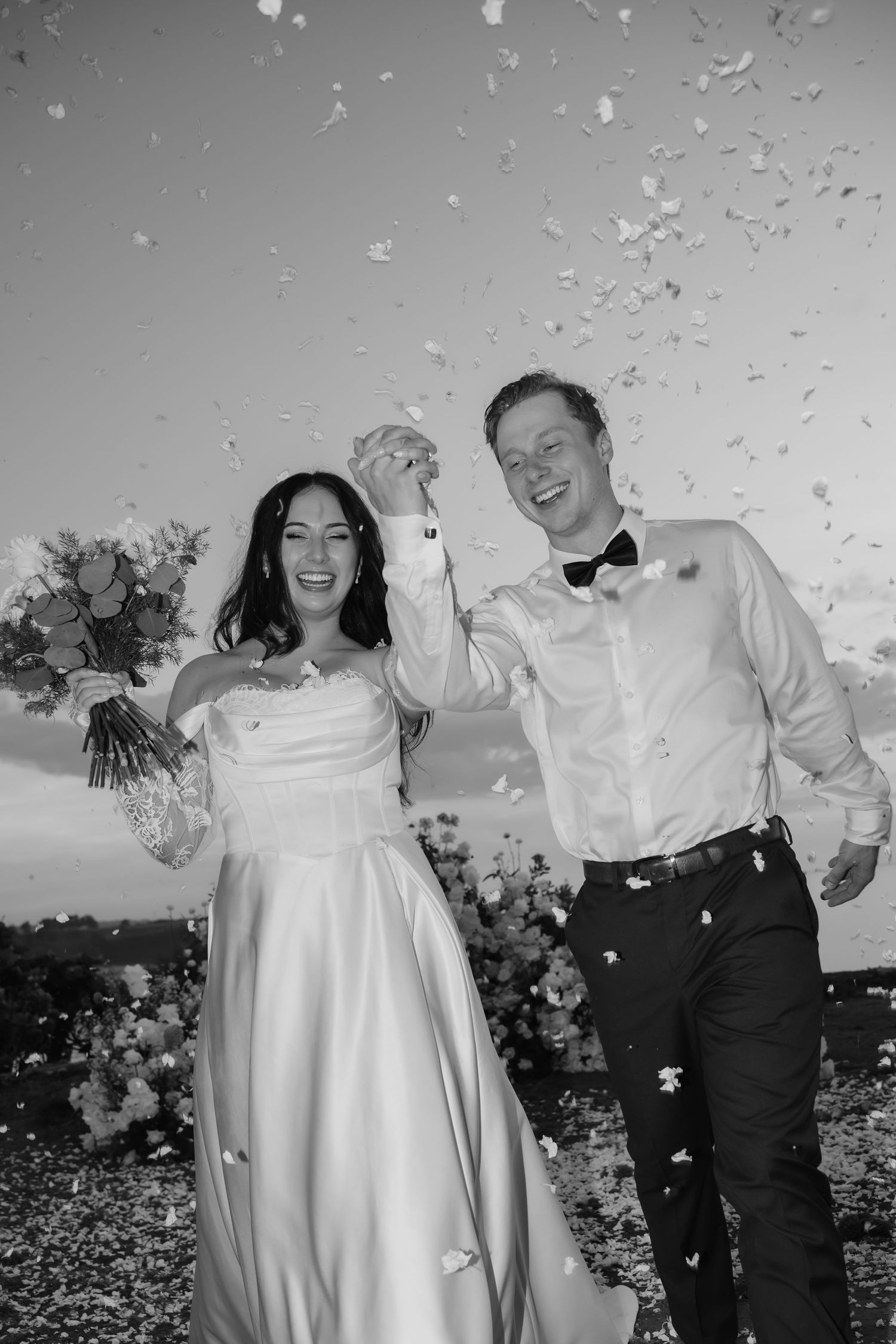 Joyful newlyweds celebrate with confetti shower in black and white wedding photo.