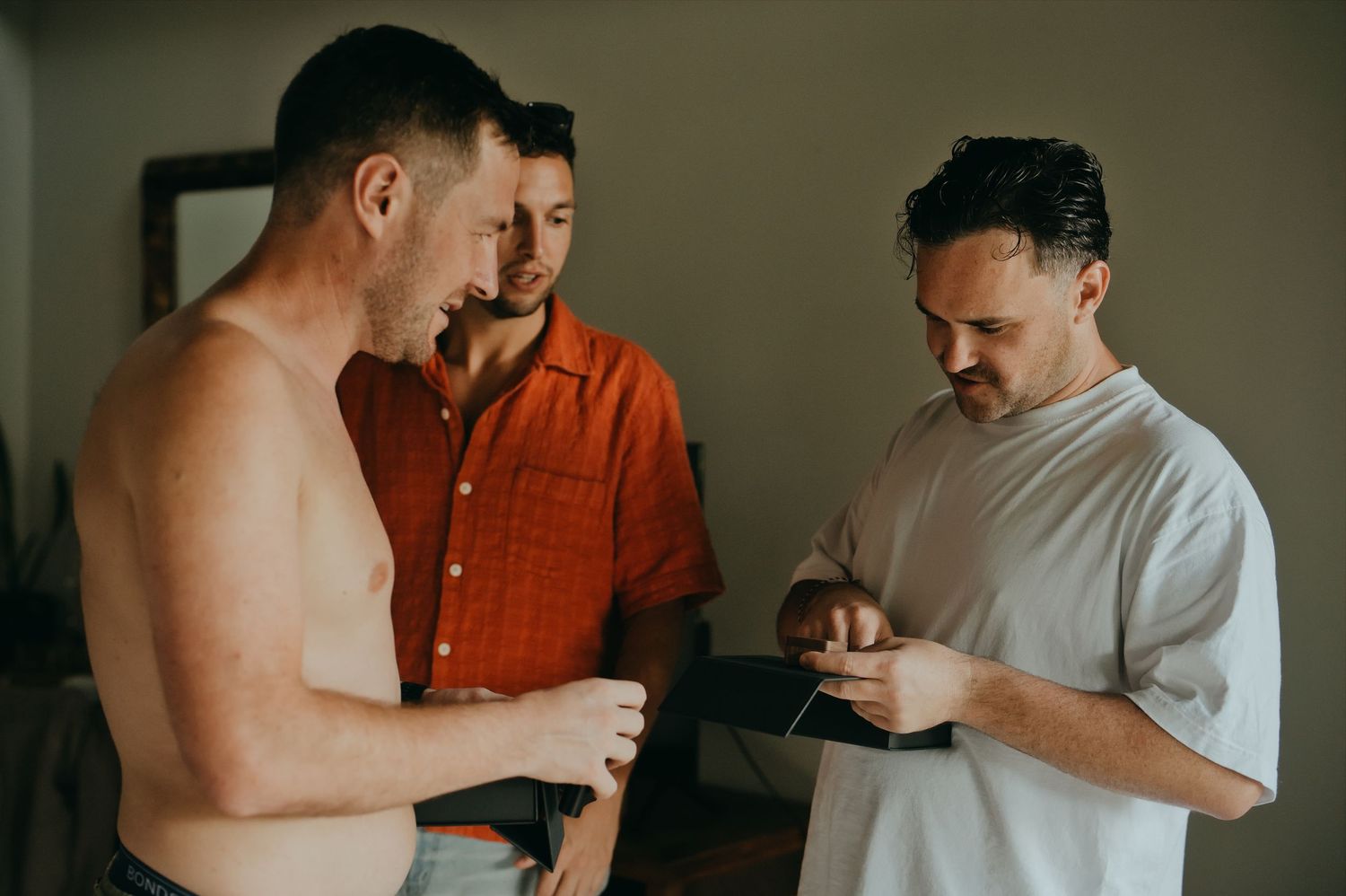 Three men getting ready together, one shirtless, sharing a lighthearted moment indoors.