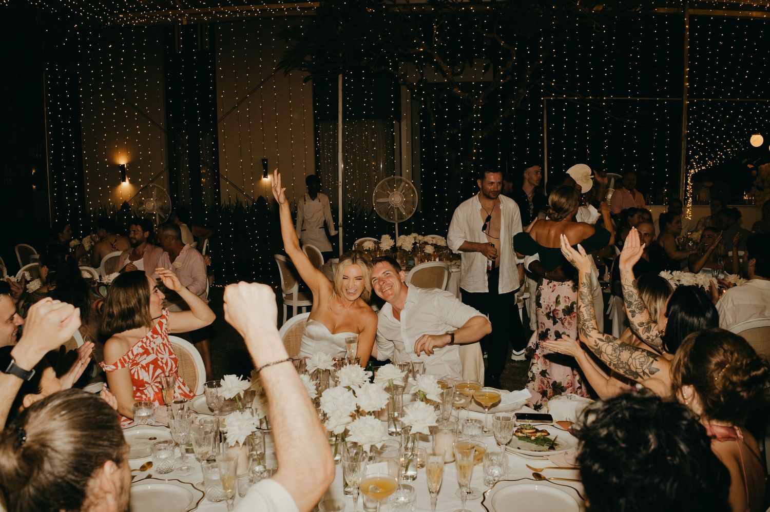 Lively wedding reception celebration with guests raising sparklers and hands at a fairy light decorated venue.