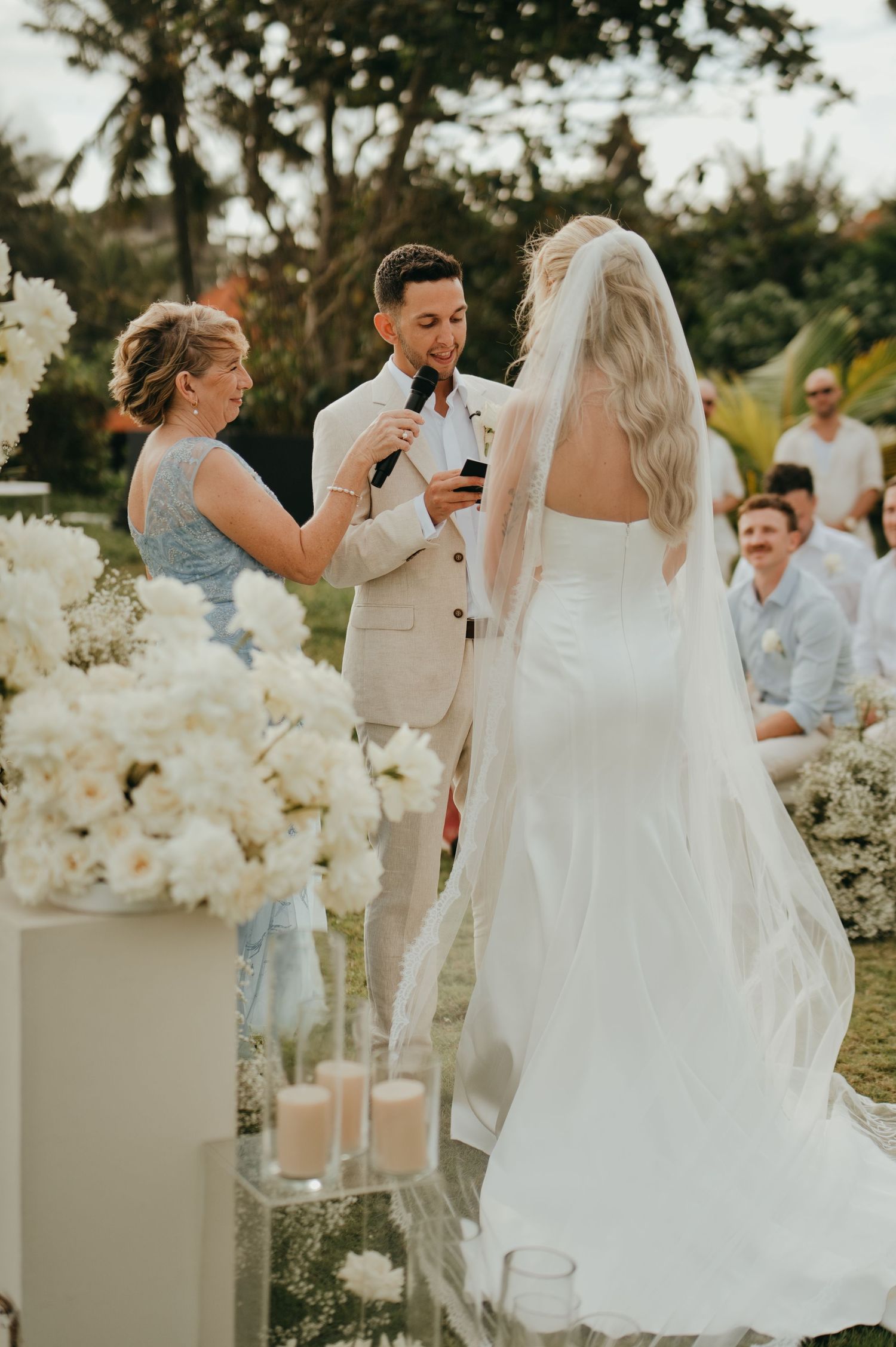 Groom reading vows into microphone while bride stands before him at elegant outdoor wedding ceremony.