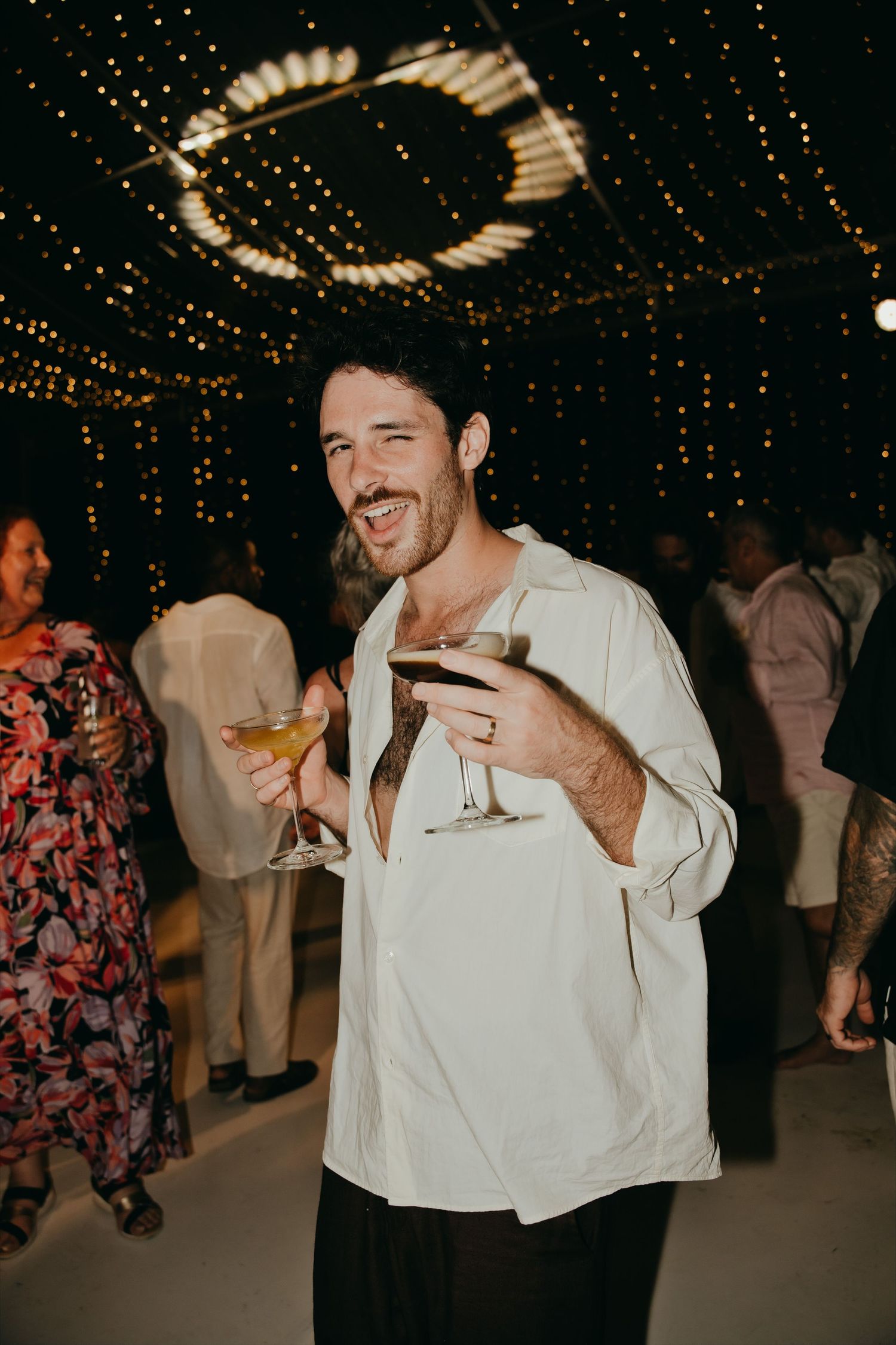Smiling male wedding guest holding two cocktail glasses under twinkling lights at a reception.