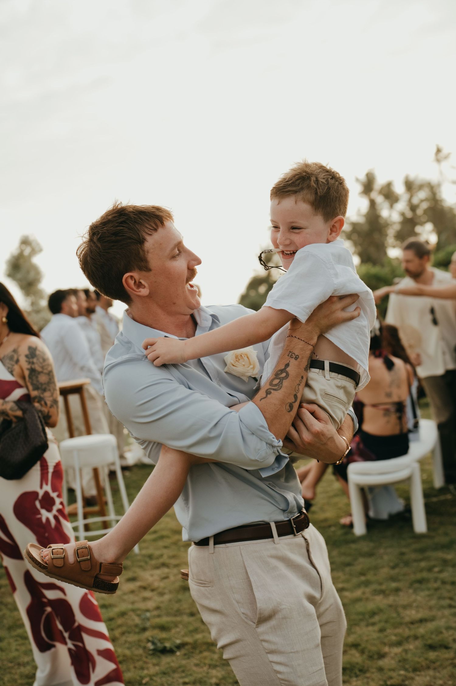 Laughing man lifts a smiling young boy at an outdoor wedding reception on a lawn.