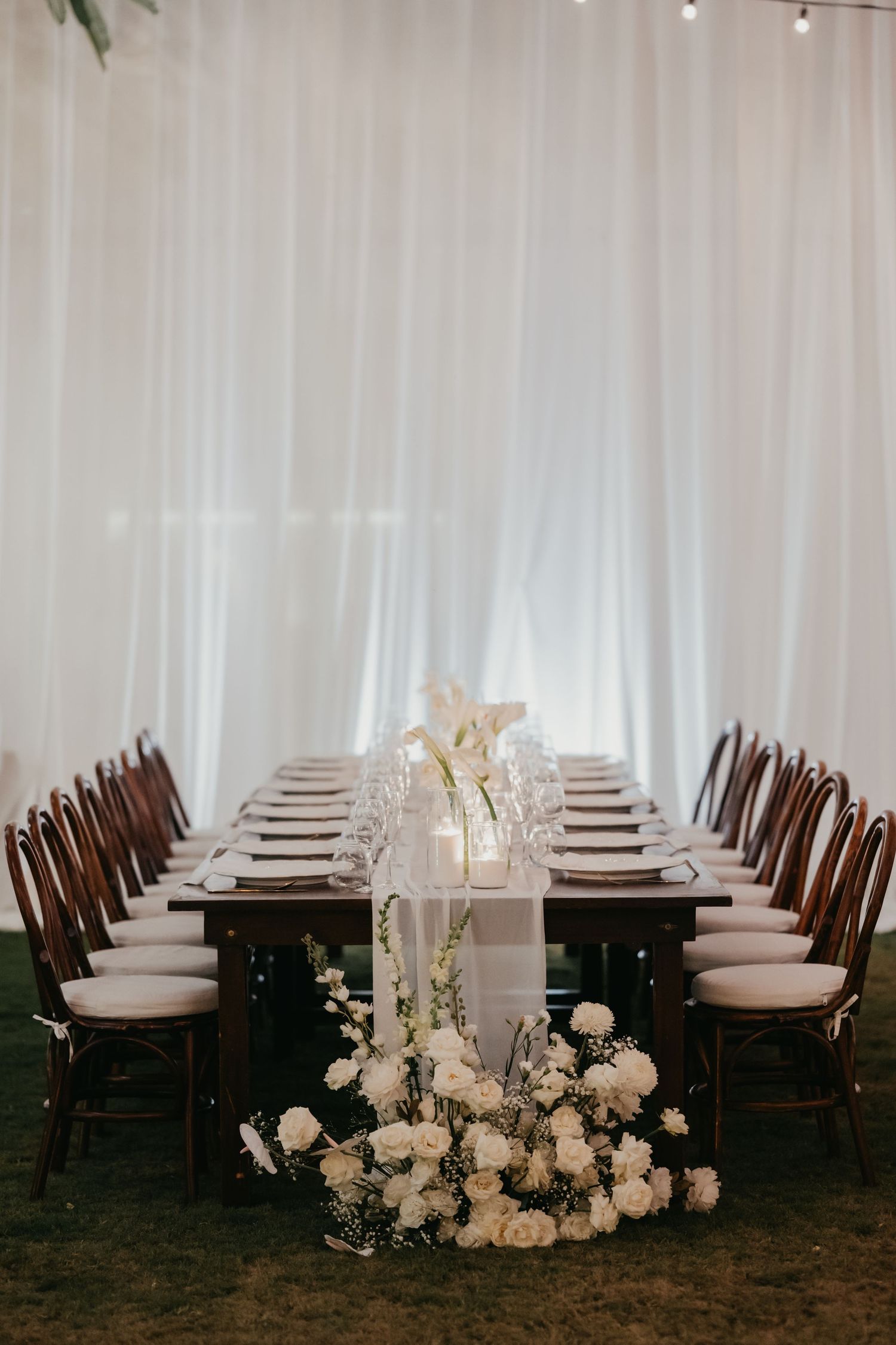 Elegant wedding reception table with white floral centerpieces and candles against white drape backdrop.
