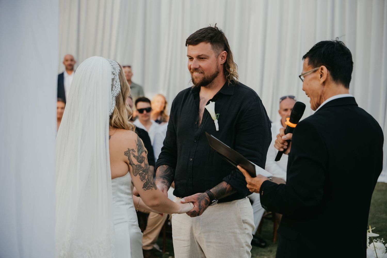 Groom with tattoos holds brides hands during outdoor wedding ceremony while officiant reads from a book.
