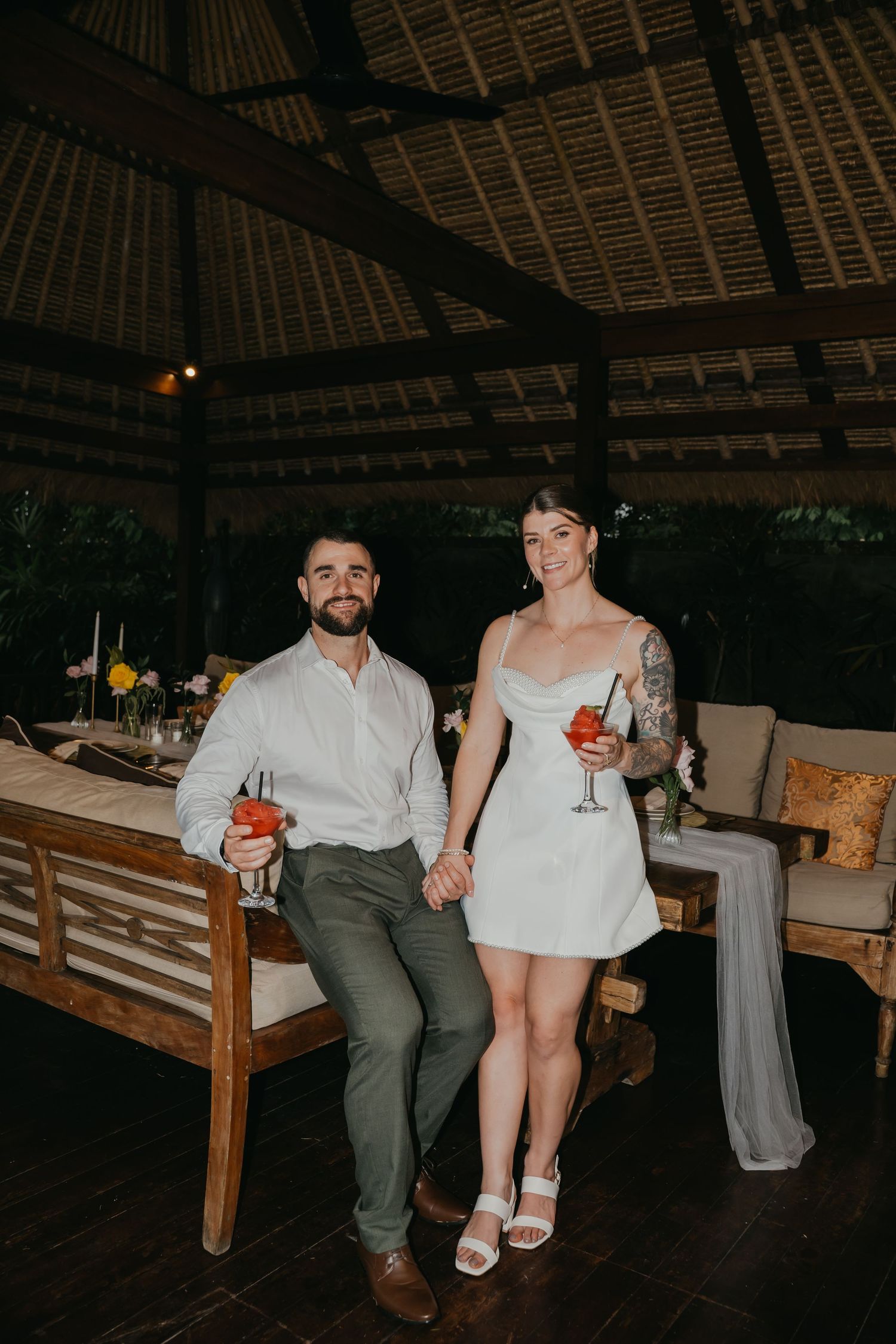 Smiling couple holding cocktails in short white dress and green trousers at reception.