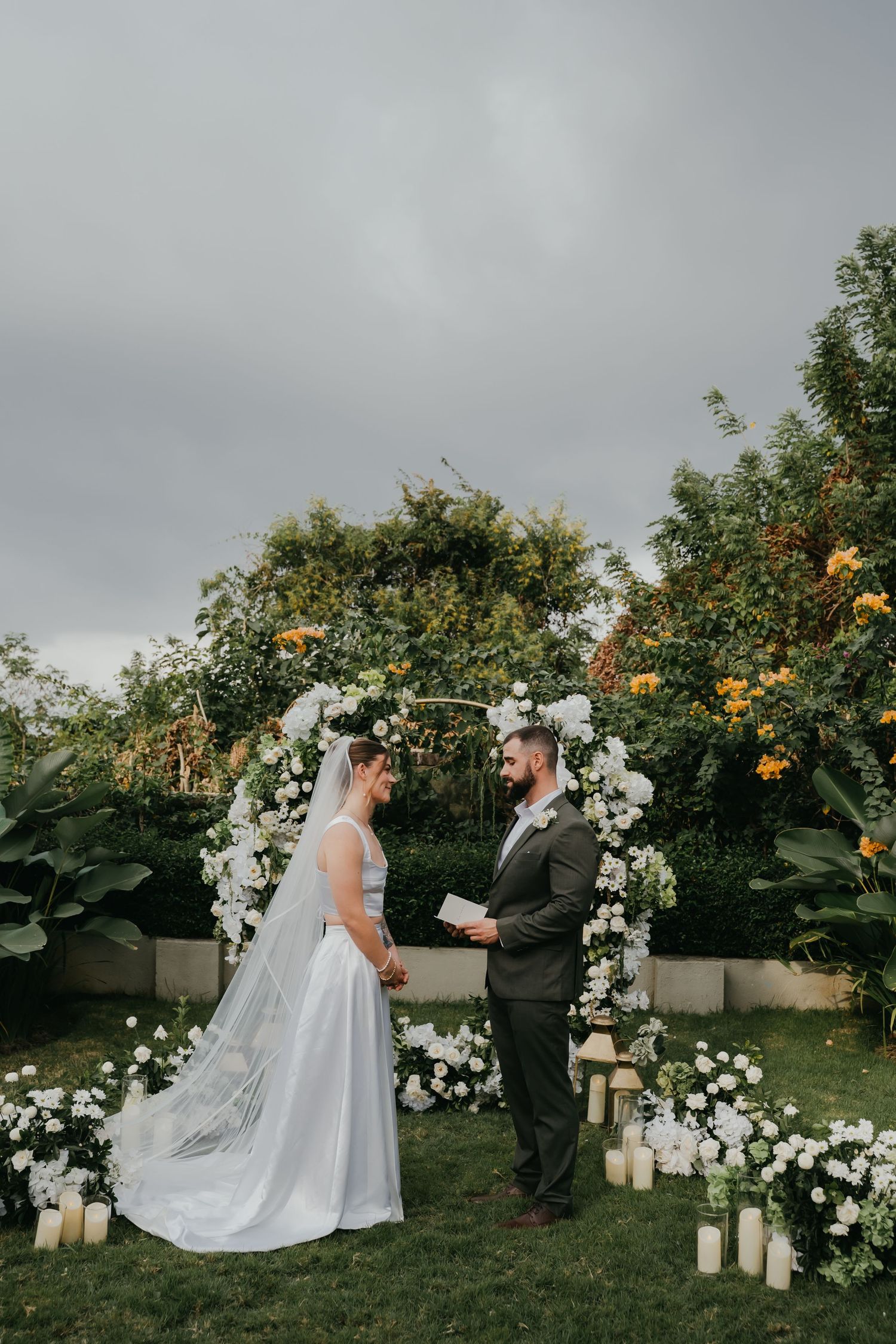 Bride and groom exchanging vows at outdoor garden wedding ceremony with white floral arch and candles.