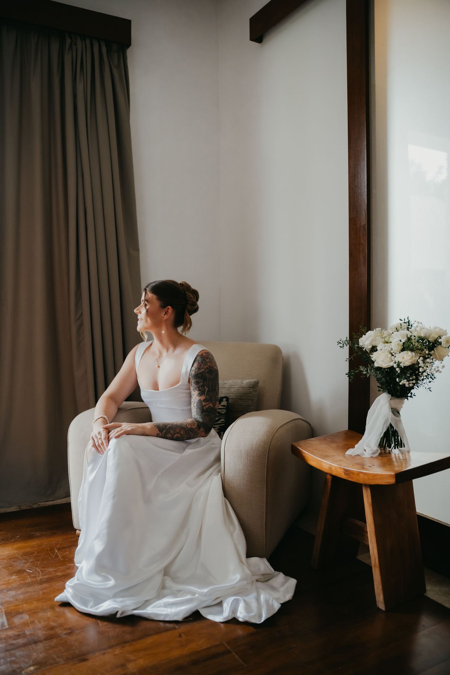 Tattooed bride in white gown sits in armchair gazing out window beside vase of white roses.