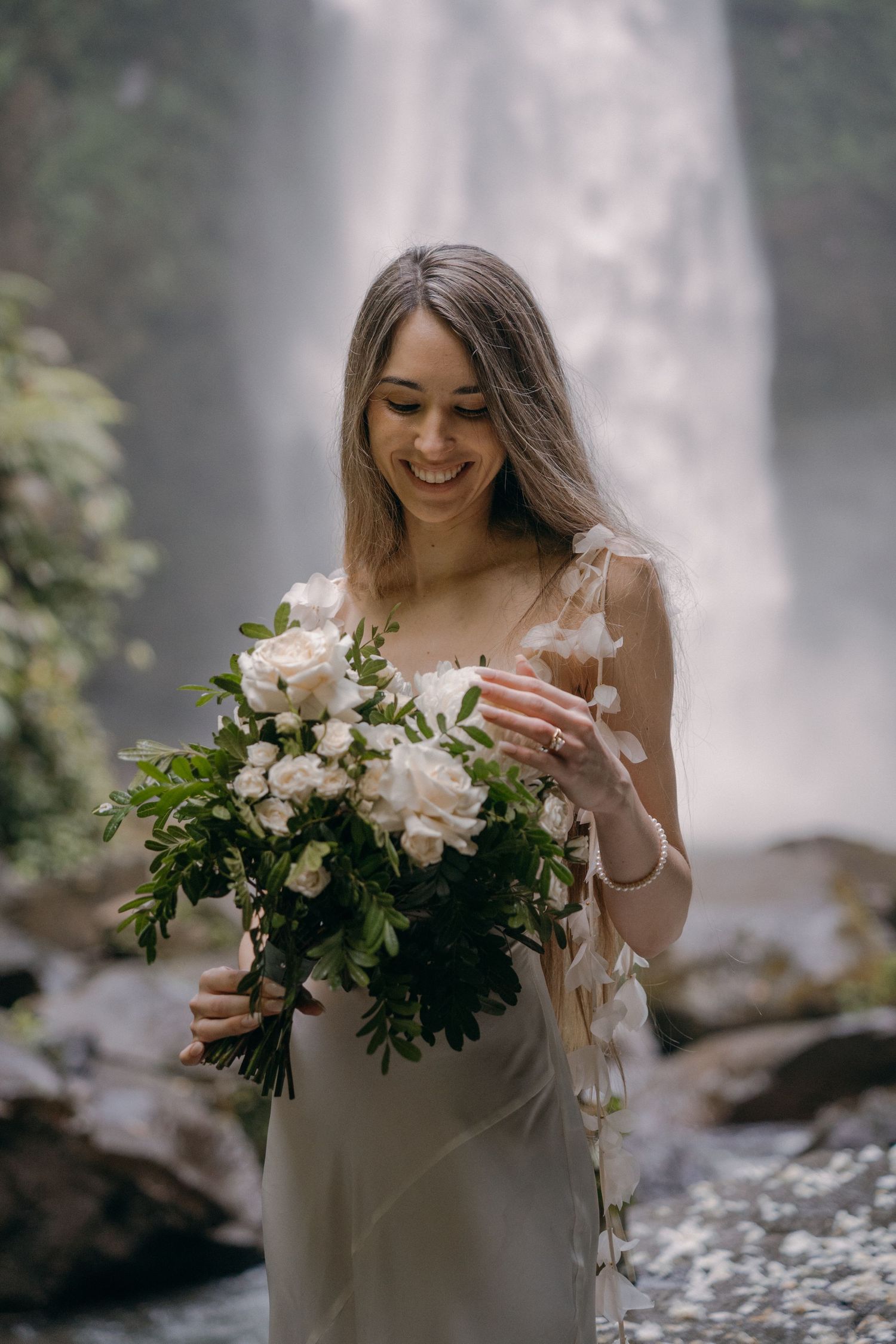 A person in a white dress holds a beautiful bouquet of white flowers in front of a misty waterfall backdrop.