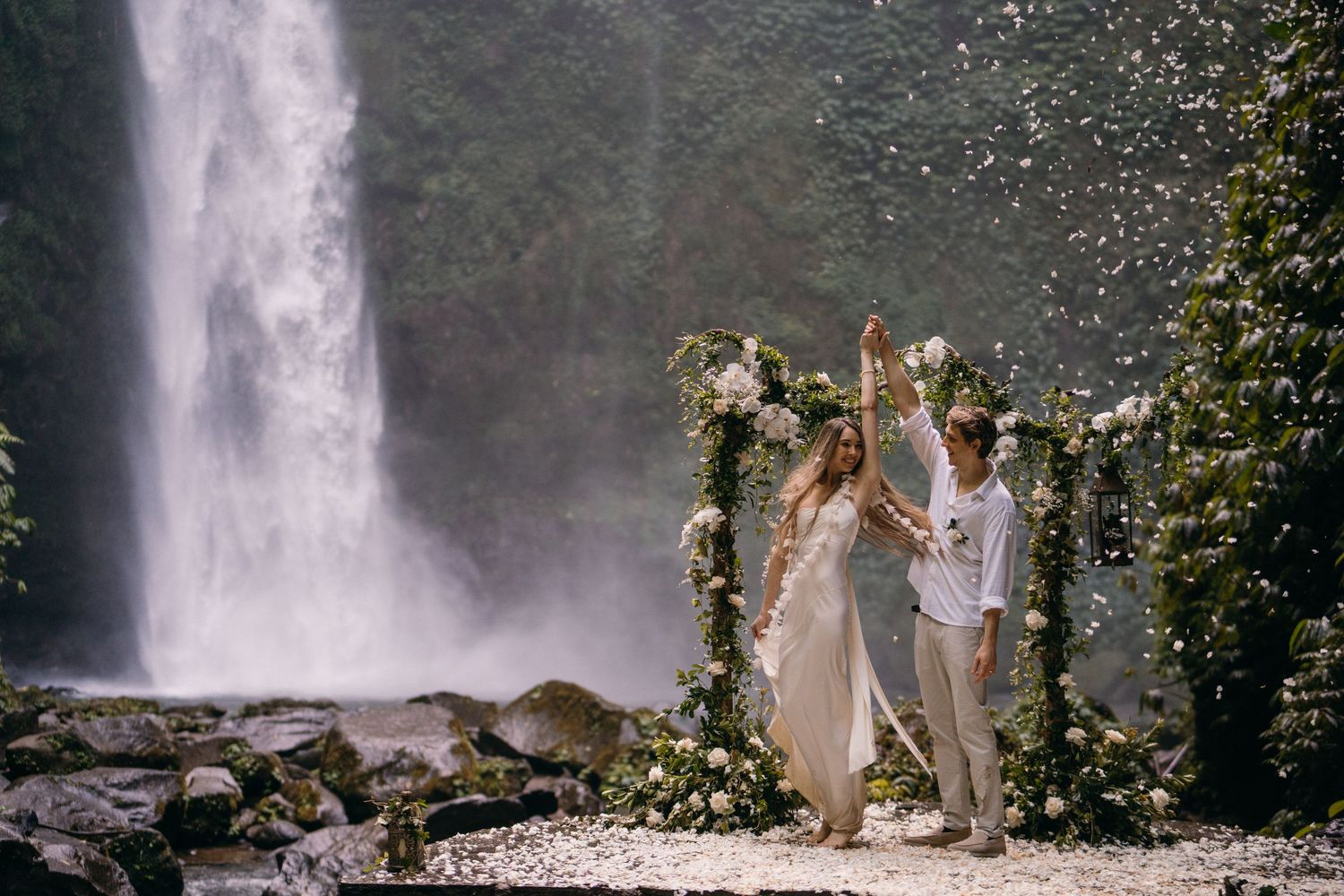 A couple stands beneath a floral arch during an intimate waterfall ceremony in a lush forest setting.