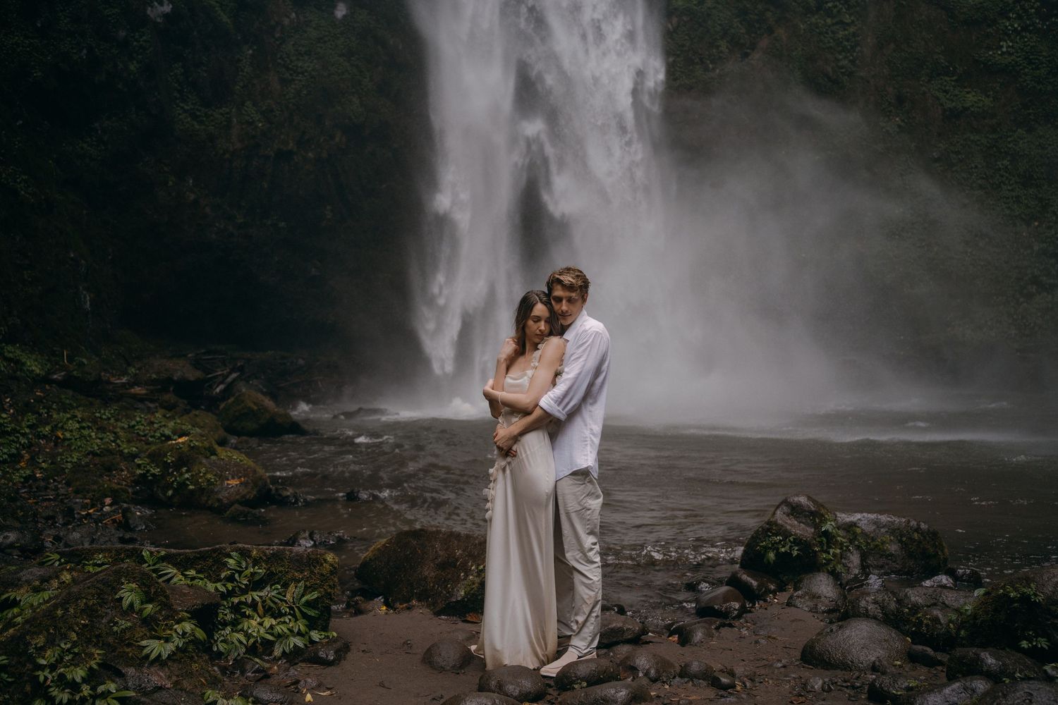 A person in a white dress stands in front of a cascading waterfall surrounded by lush greenery.
