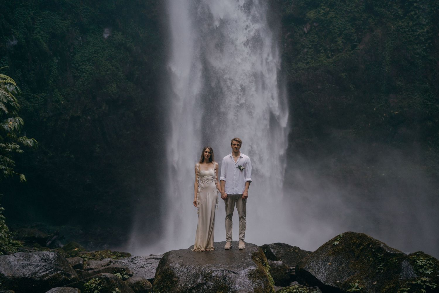 A couple poses in front of a majestic waterfall in a lush forest setting for romantic engagement photos.