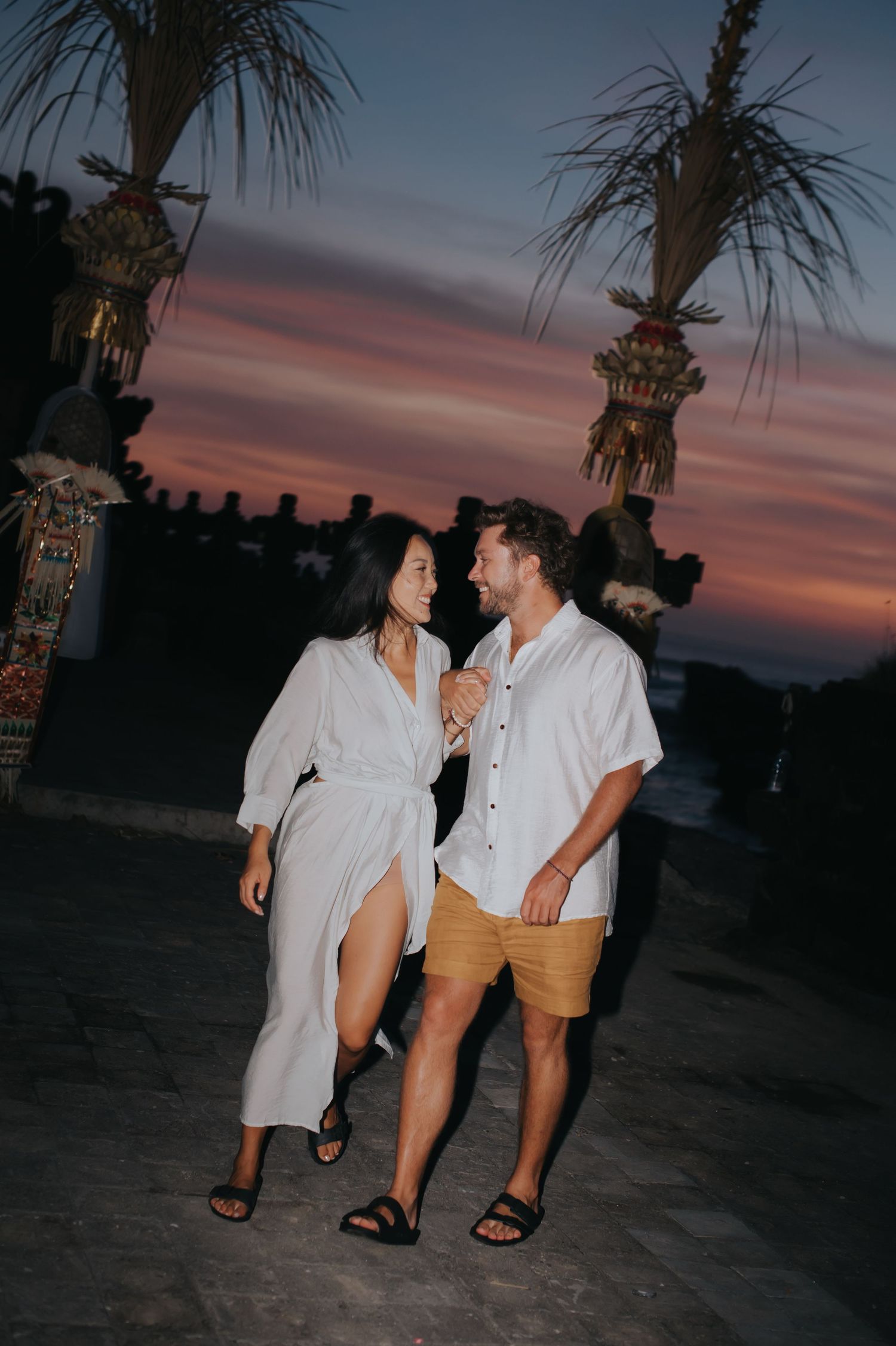 A couple in summer attire enjoying a romantic moment at sunset with palm trees and warm sky in the background.