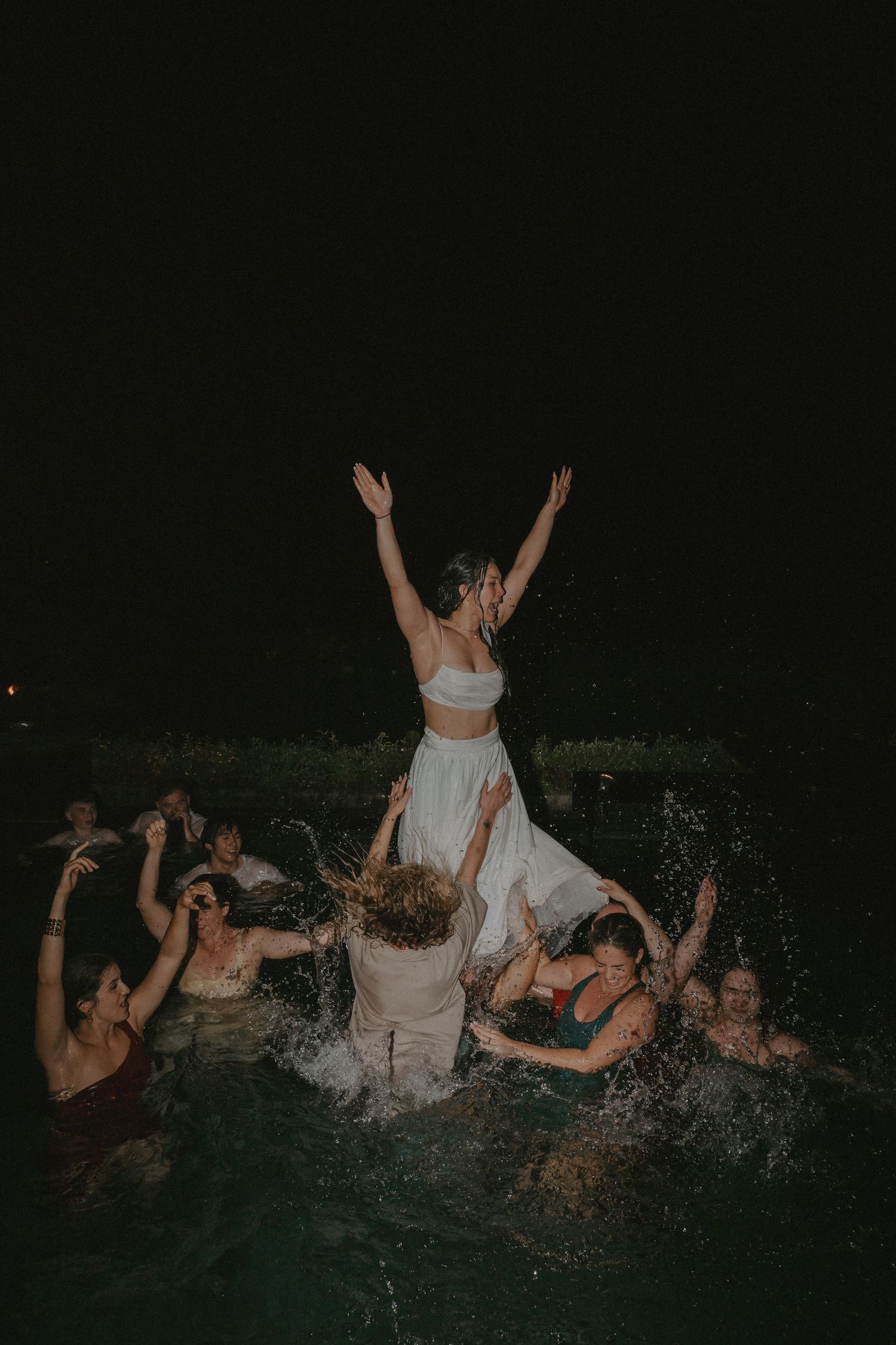 Group of people splashing and playing in dark water at night, creating dramatic silhouettes against the black background.