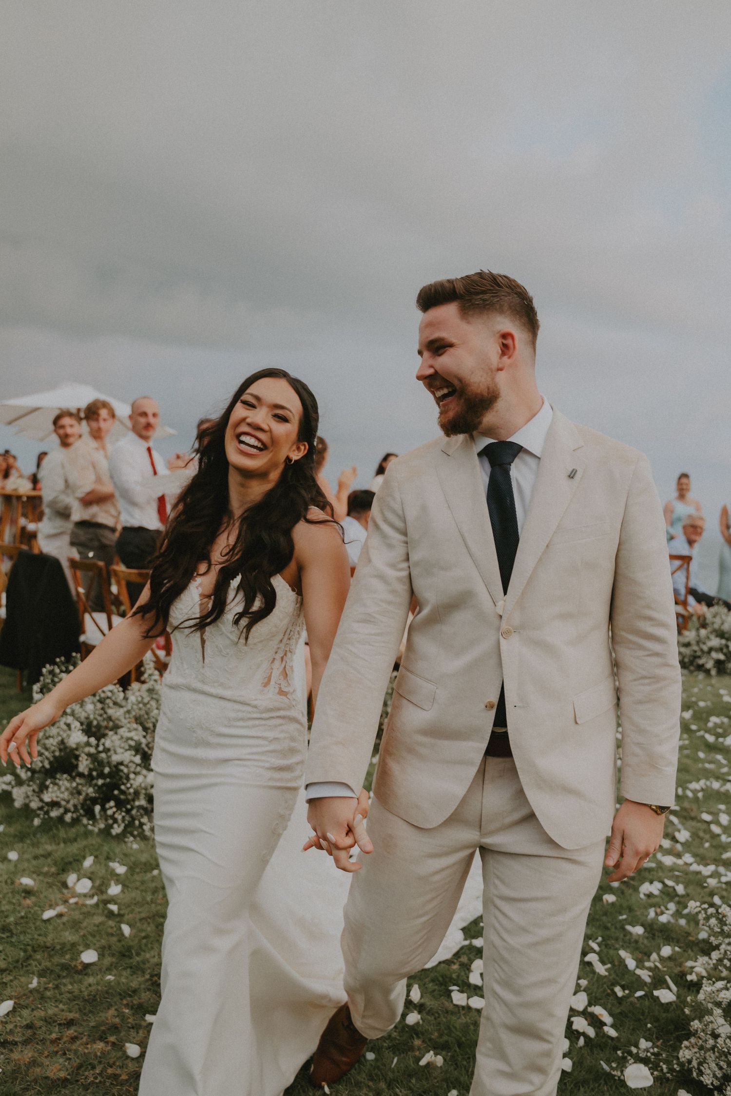 A joyful couple in wedding attire walk hand in hand through a flower-filled meadow under cloudy skies.