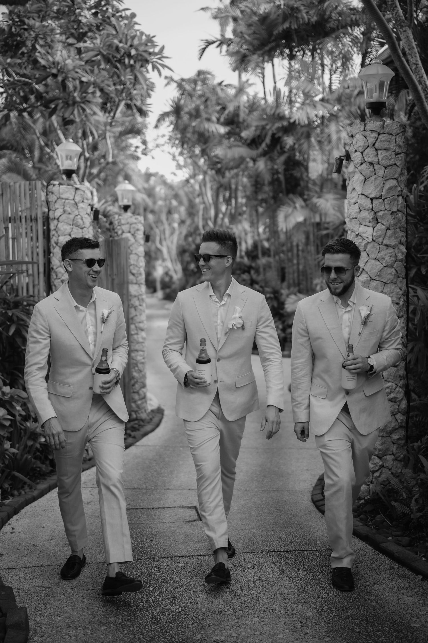 Groomsmen in matching light suits walk together along garden path in black and white photo.