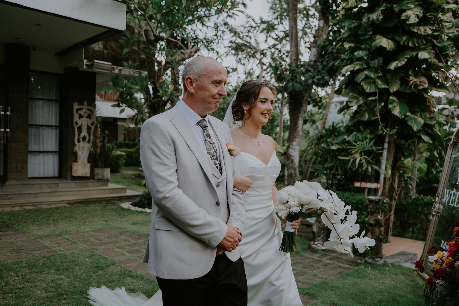 Father walking bride down outdoor aisle in white wedding gown surrounded by tropical garden setting.