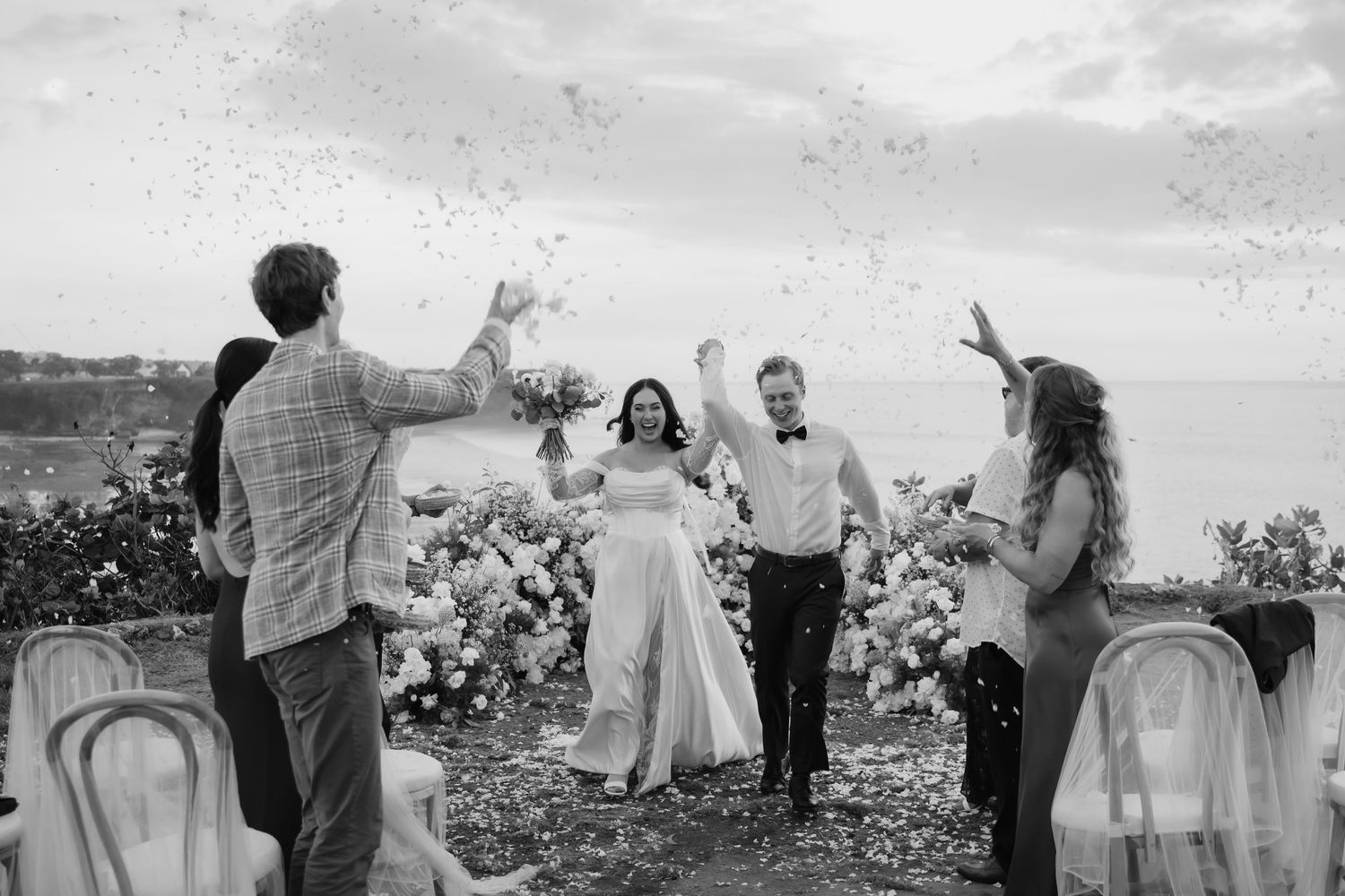 Bride and groom celebrate with confetti as guests cheer during outdoor wedding ceremony exit.