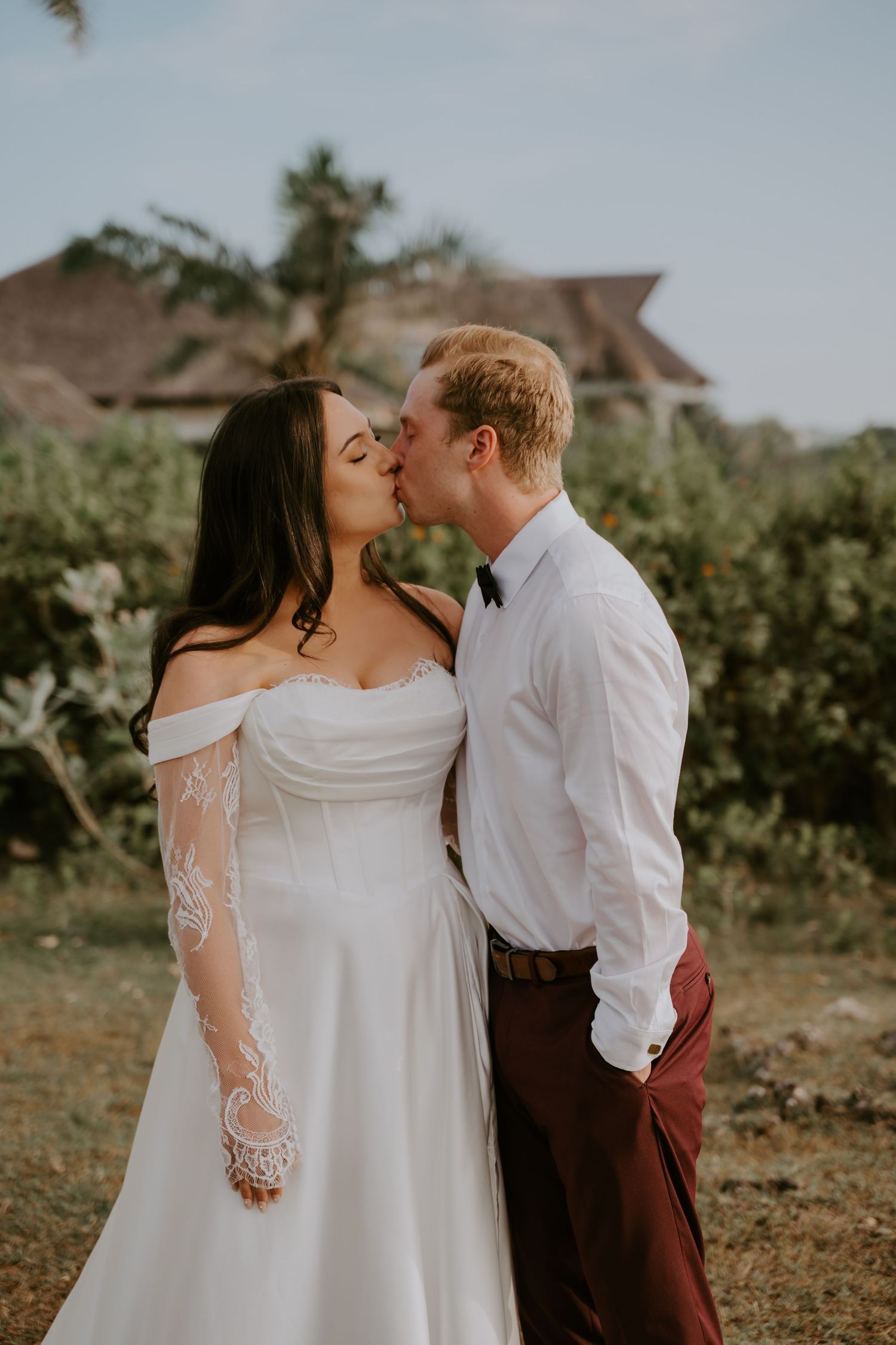 Bride and groom share a romantic kiss outdoors, she wearing an off-shoulder lace wedding gown, he in white shirt and burgundy pants.