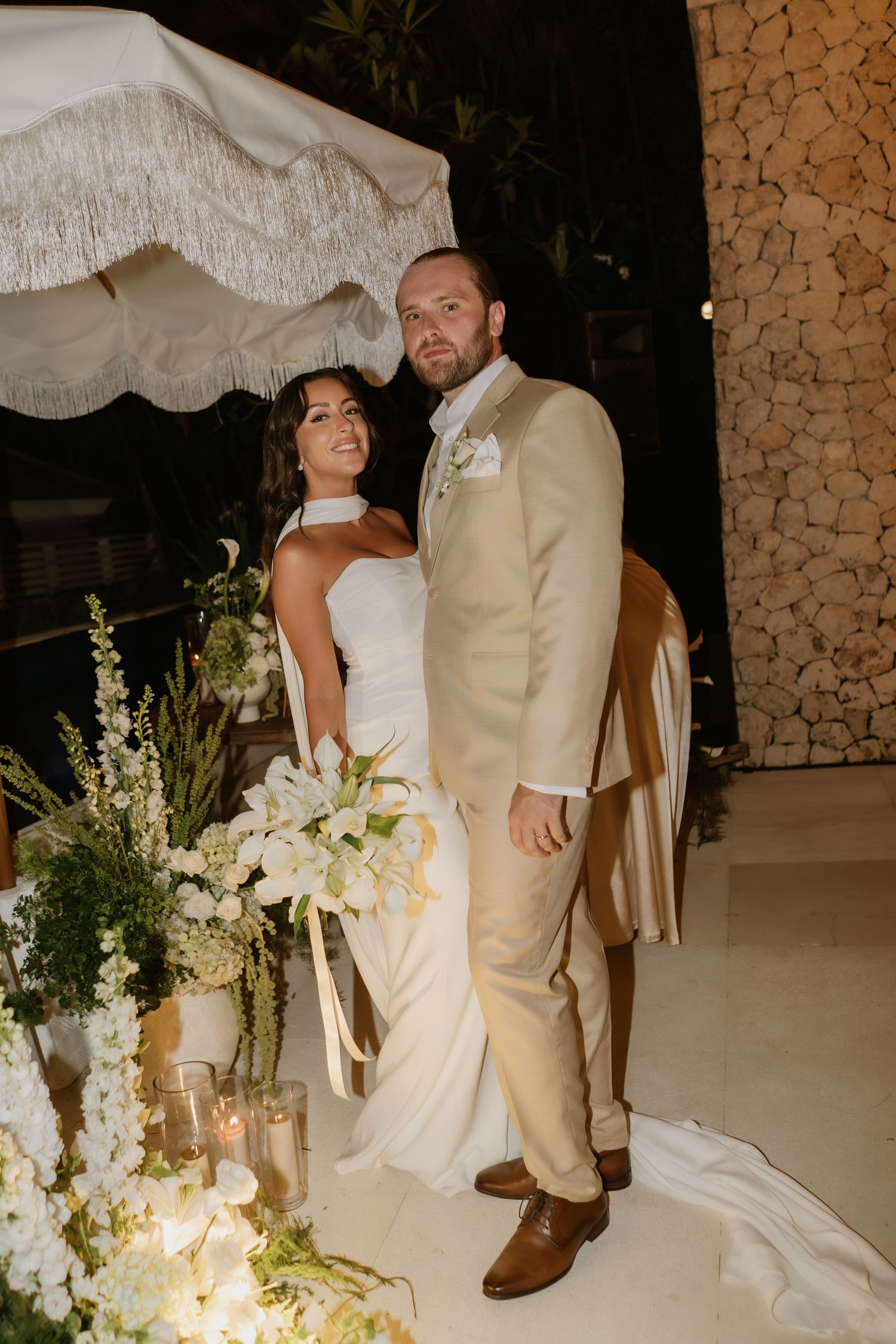 Bride and groom pose together at night beside lush white floral arrangements under fringed umbrella.