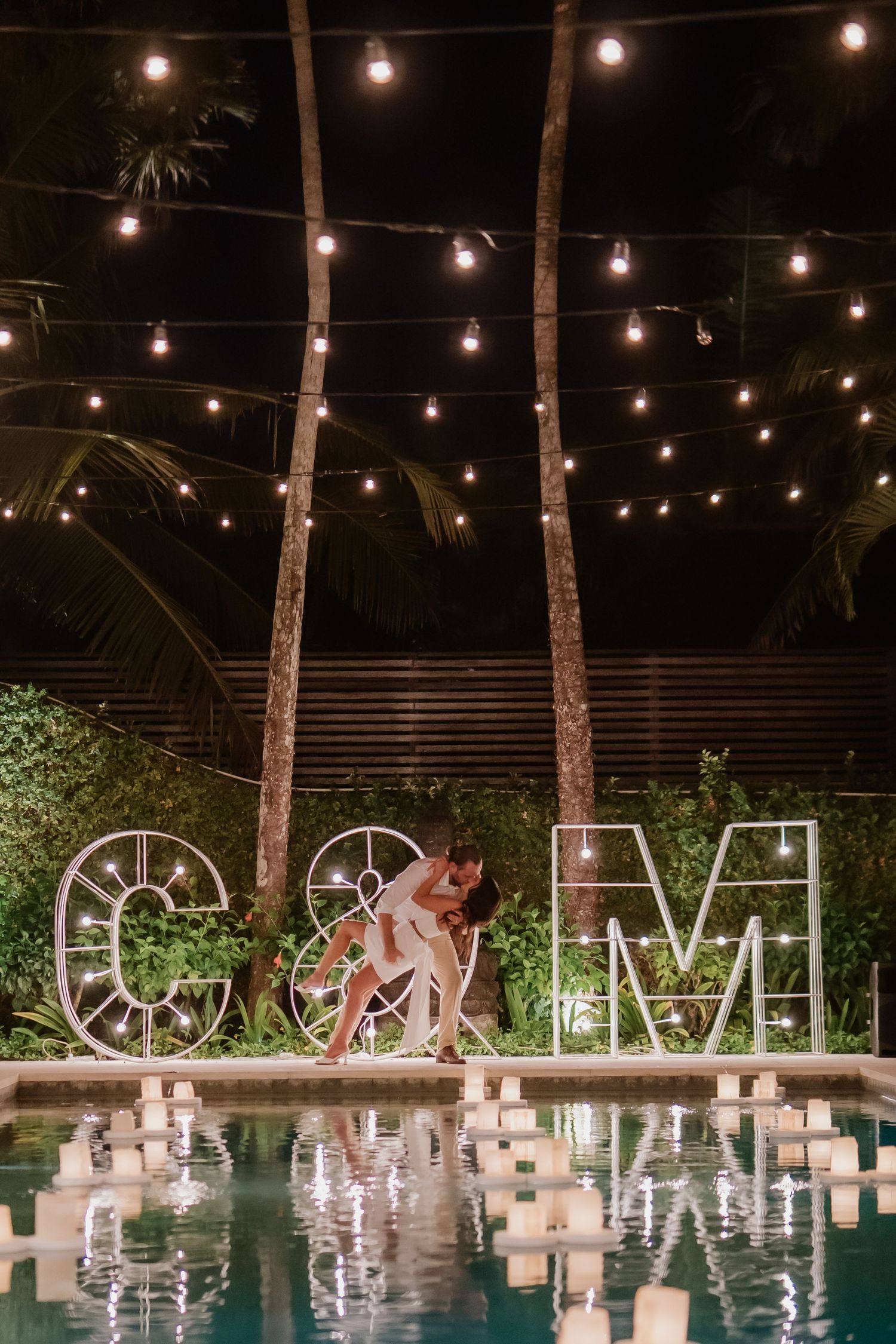 Couple embraces by illuminated letter marquee signs beside candle-lined pool under string lights and palm trees at night wedding.