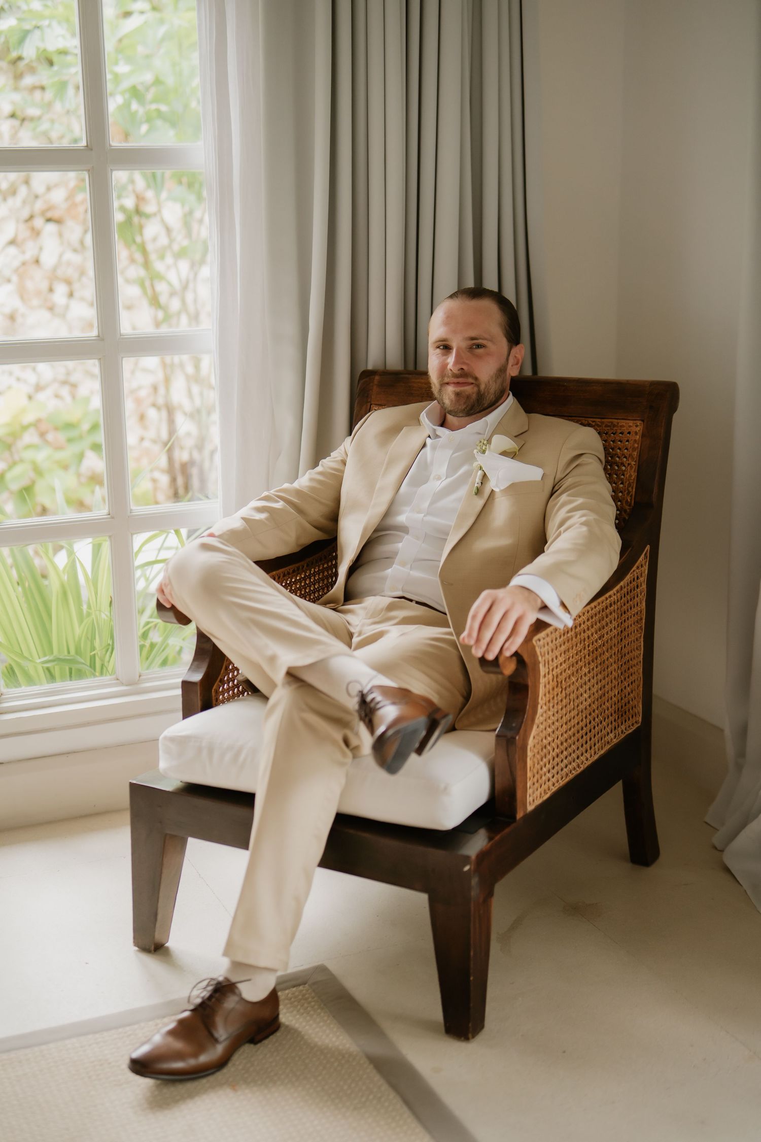 Groom in beige suit sitting relaxed in wooden rattan chair by window with white boutonniere.
