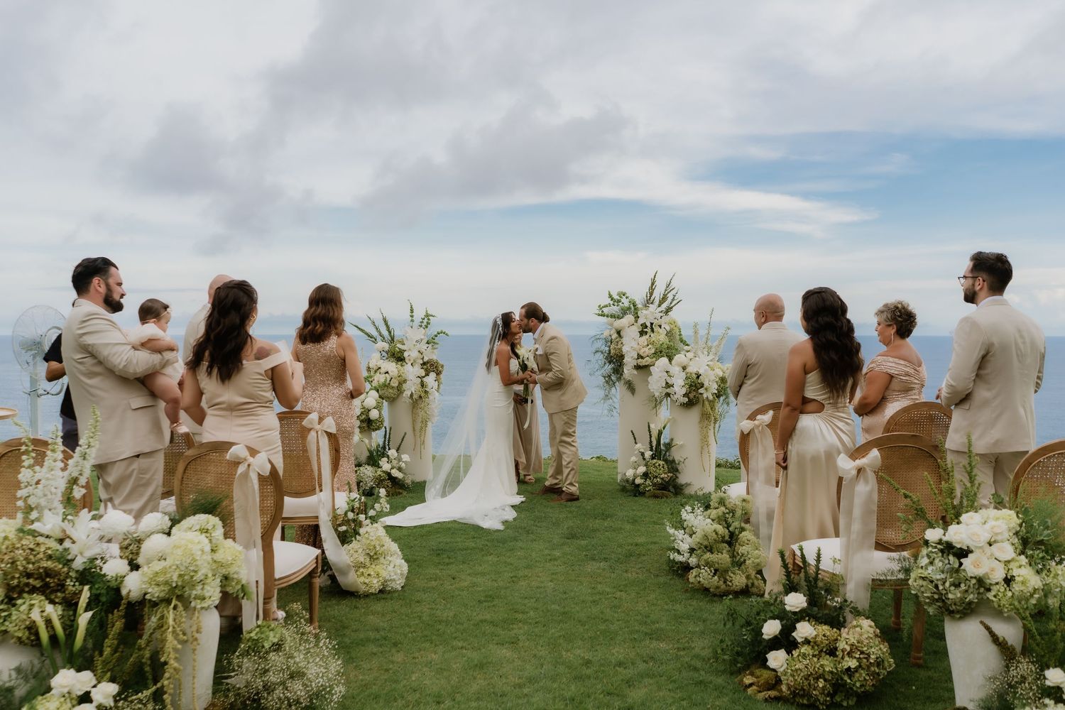Couple kisses at outdoor wedding ceremony with white floral arrangements and ocean backdrop.