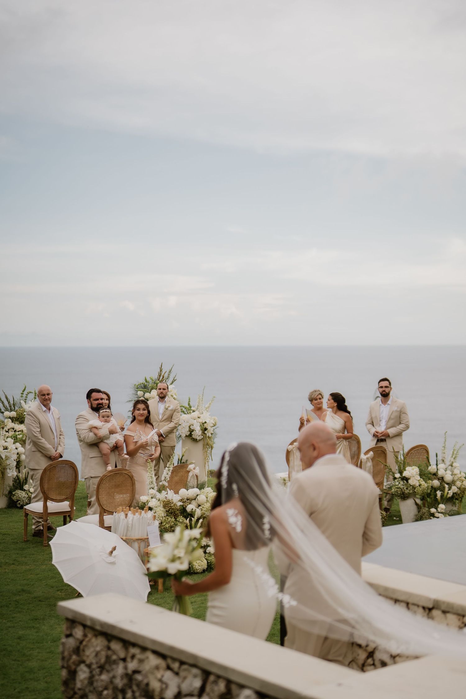Bride and groom exchange vows at an oceanfront wedding ceremony with white floral arch and seated guests.