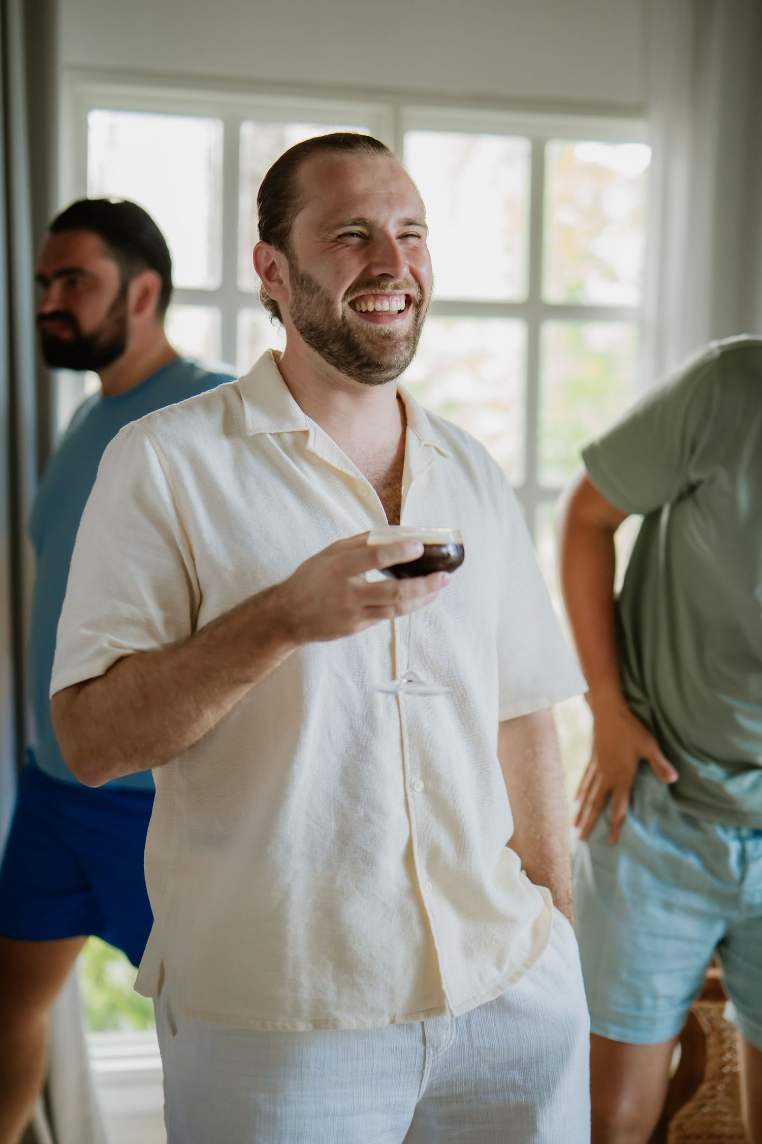 Smiling groom holds drink with groomsmen during casual wedding day preparations.