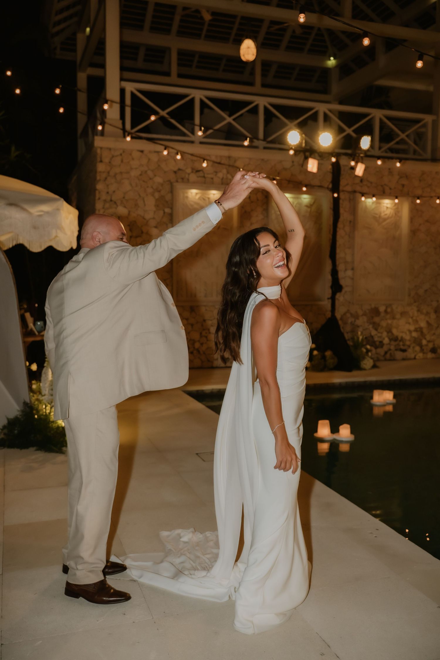 Groom twirls smiling bride in white gown beside illuminated pool at outdoor wedding reception at night.