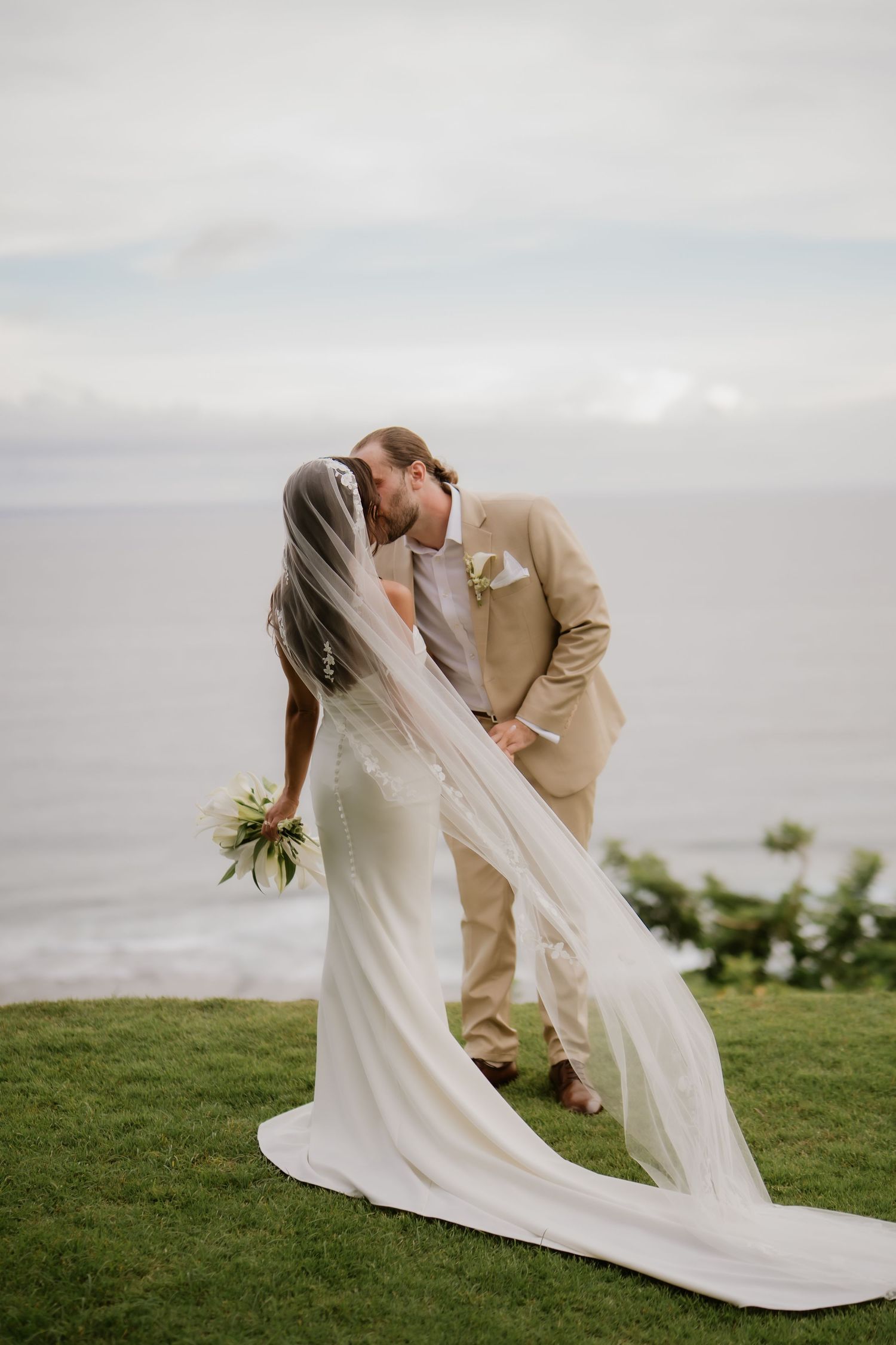 Bride and groom sharing first kiss on green lawn with dramatic ocean horizon in background.