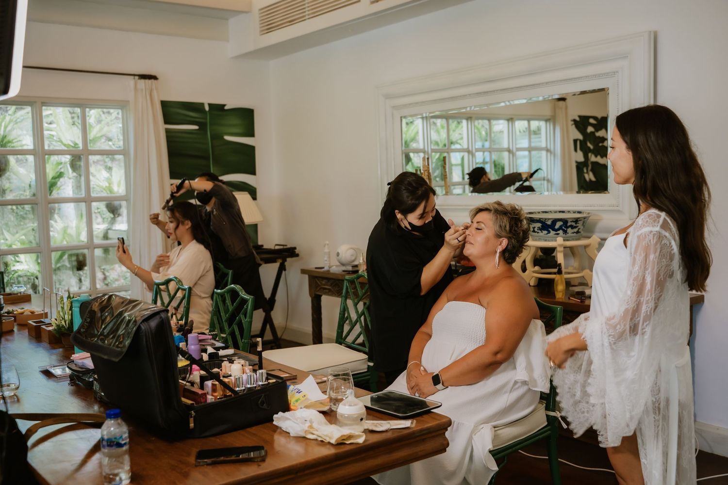 Bride getting makeup done by artist while bridesmaids prepare in bright, tropical wedding venue room.