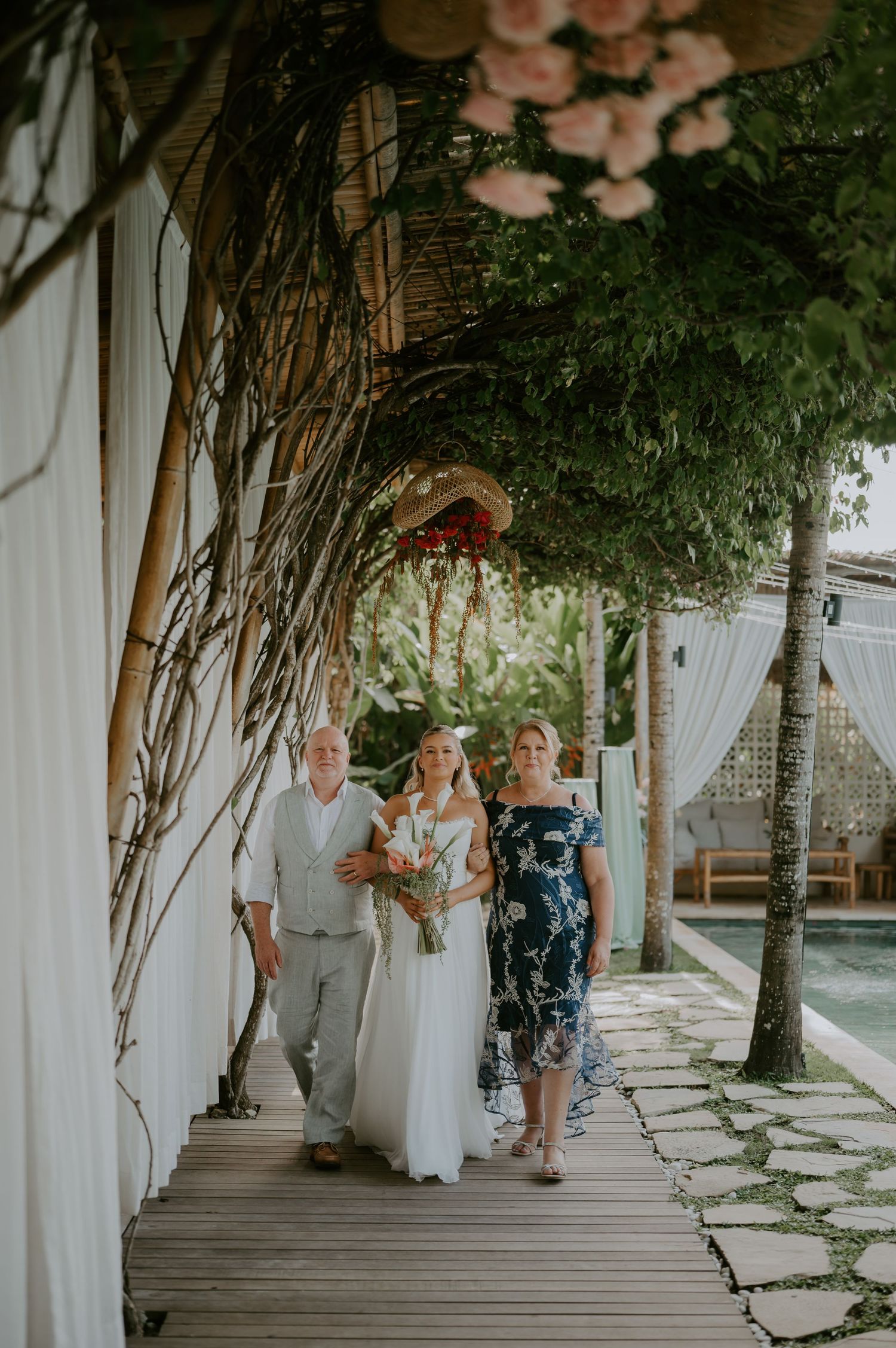Bride walking down bamboo-lined aisle with parents at tropical outdoor wedding venue.