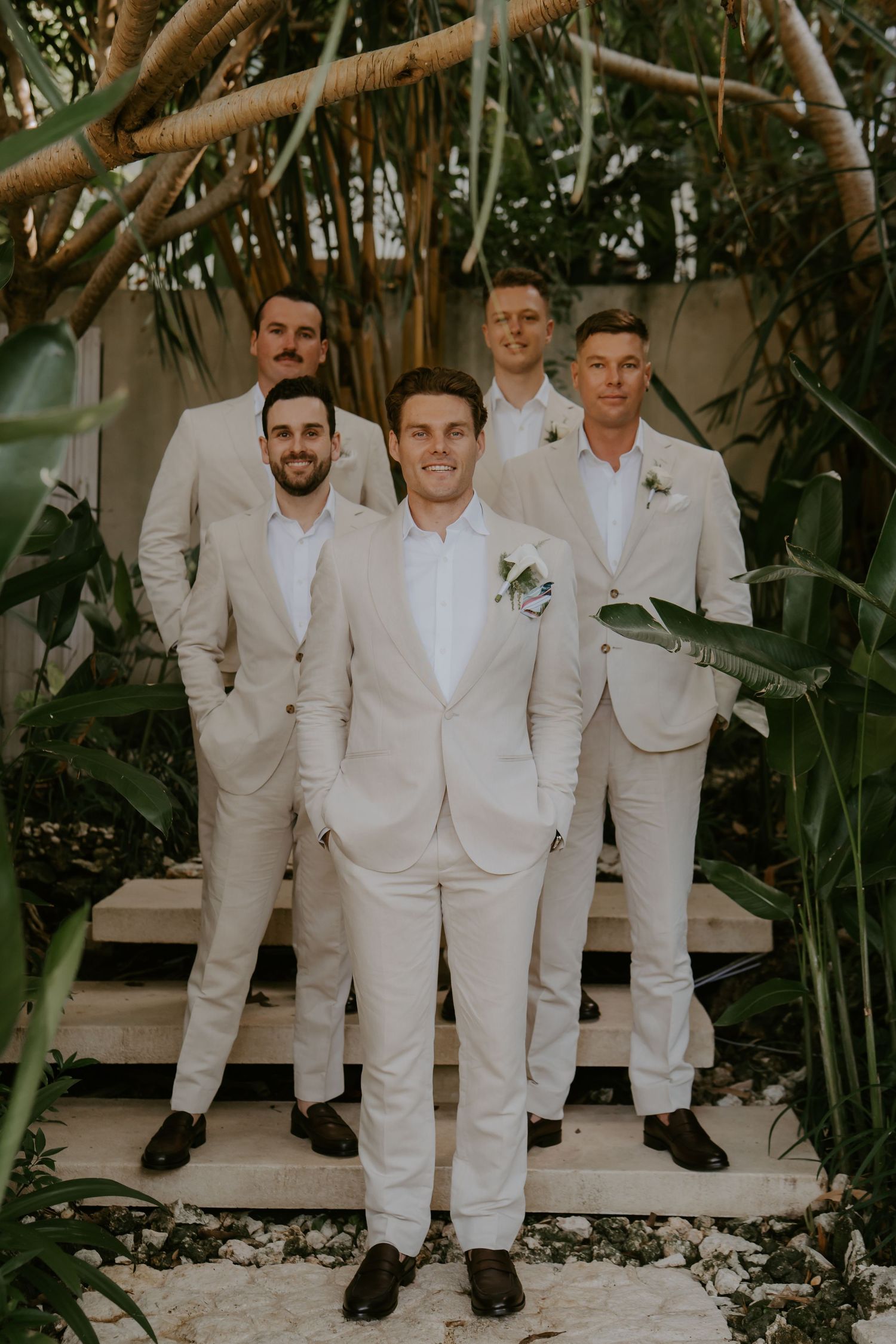 Groom in cream suit stands confidently with four groomsmen on stone steps surrounded by tropical foliage.