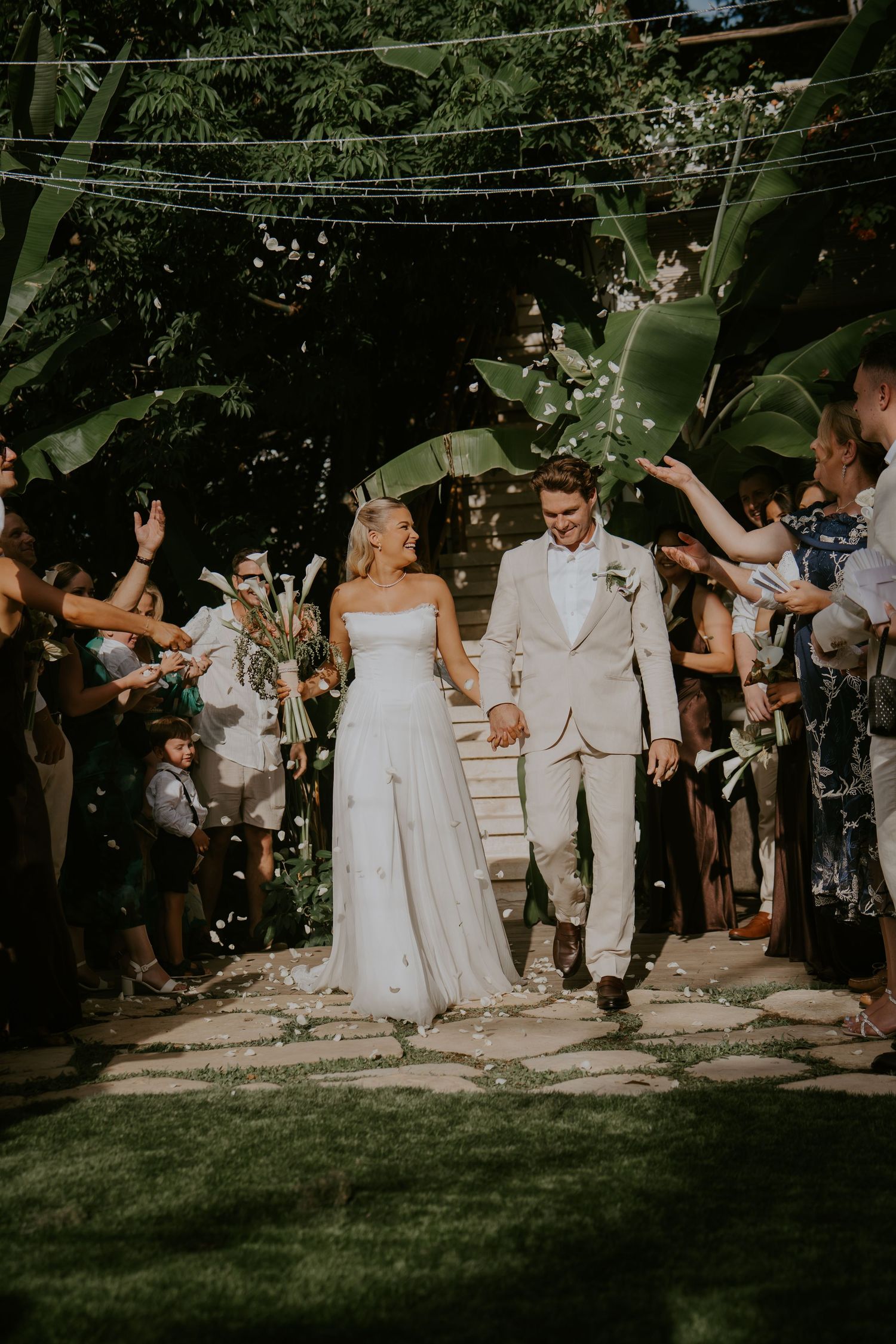 Newlyweds walk through confetti shower surrounded by cheering guests in lush garden setting.