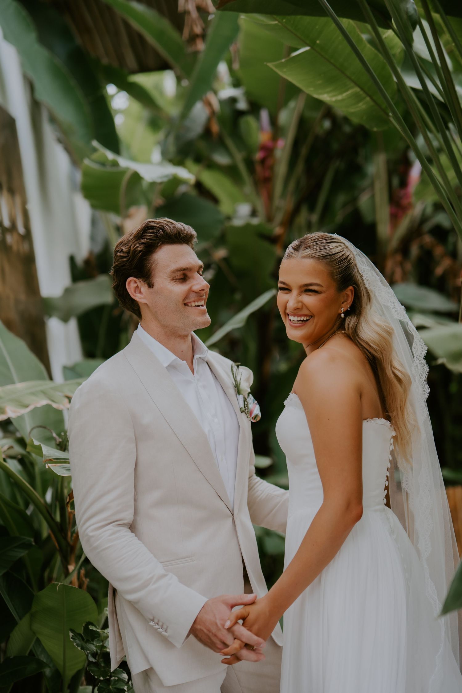 Smiling bride and groom holding hands among lush tropical greenery after ceremony.