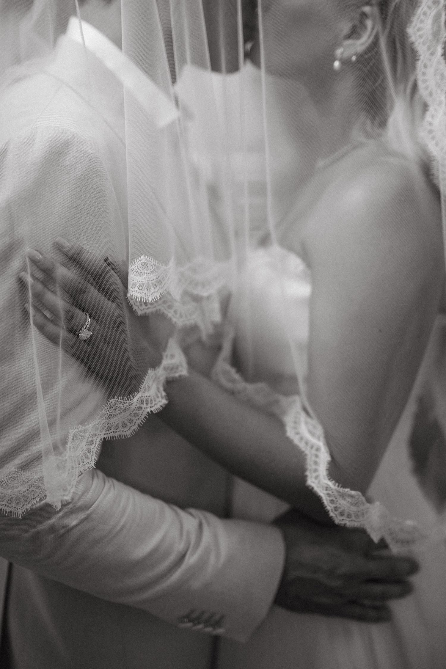 Black and white close-up of bride and groom embracing under delicate lace-trimmed veil.
