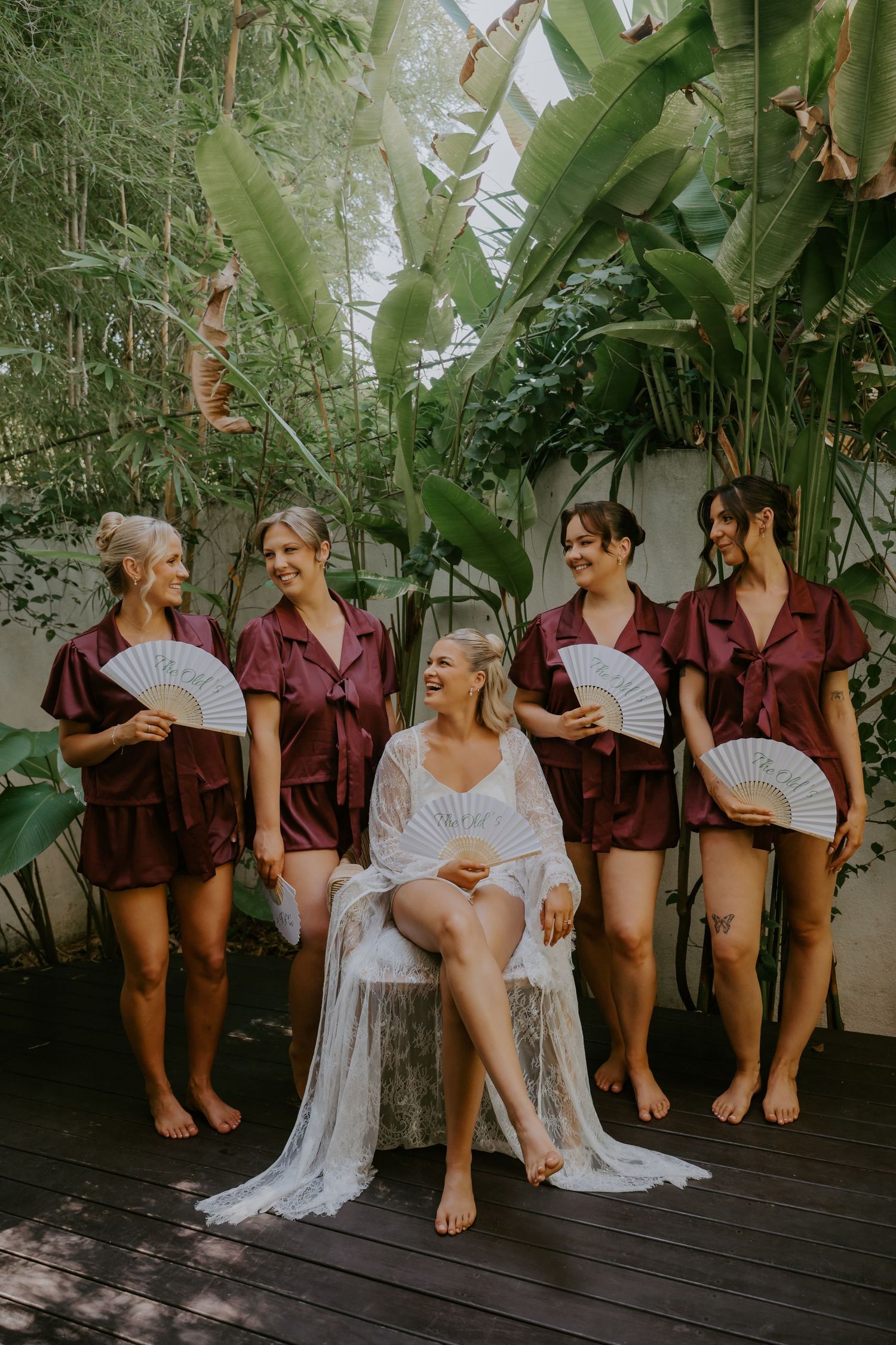 Bride in white lace robe seated on wooden stump with four bridesmaids in burgundy satin sets holding hand fans.