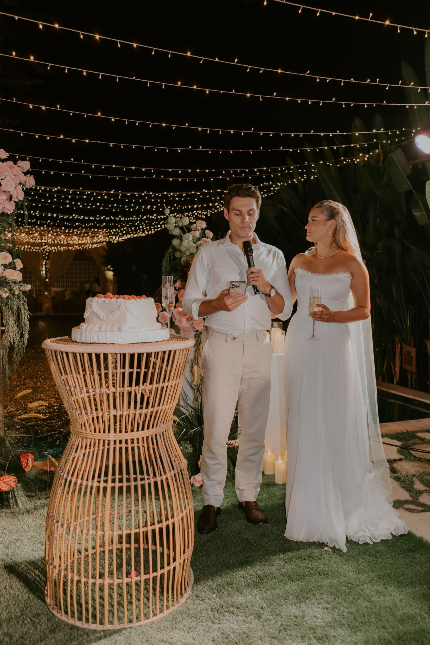 Bride and groom stand by wicker cake stand under fairy lights at outdoor night wedding reception.