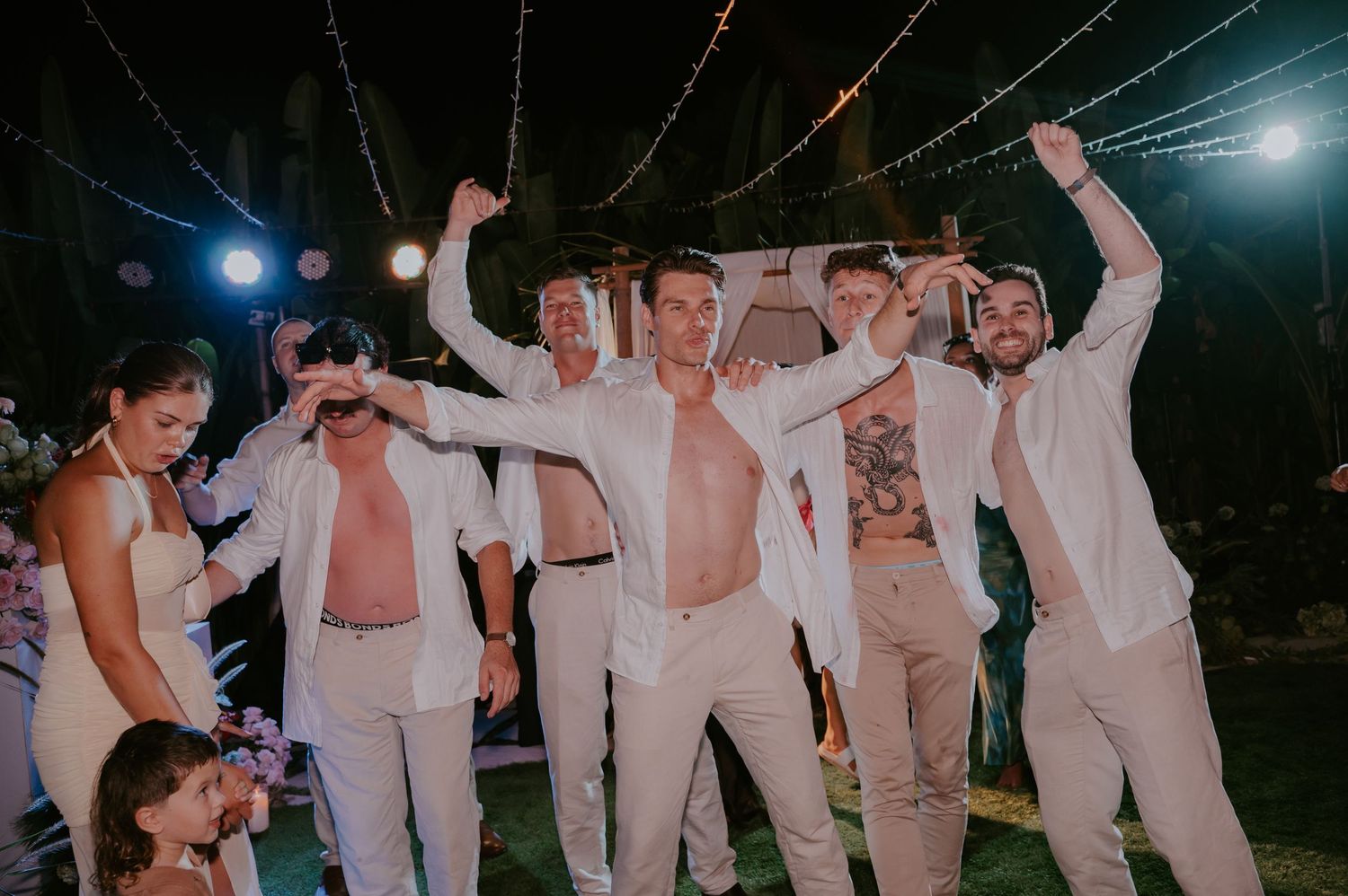Groomsmen celebrate with shirts open and arms raised under string lights at an energetic outdoor wedding reception.