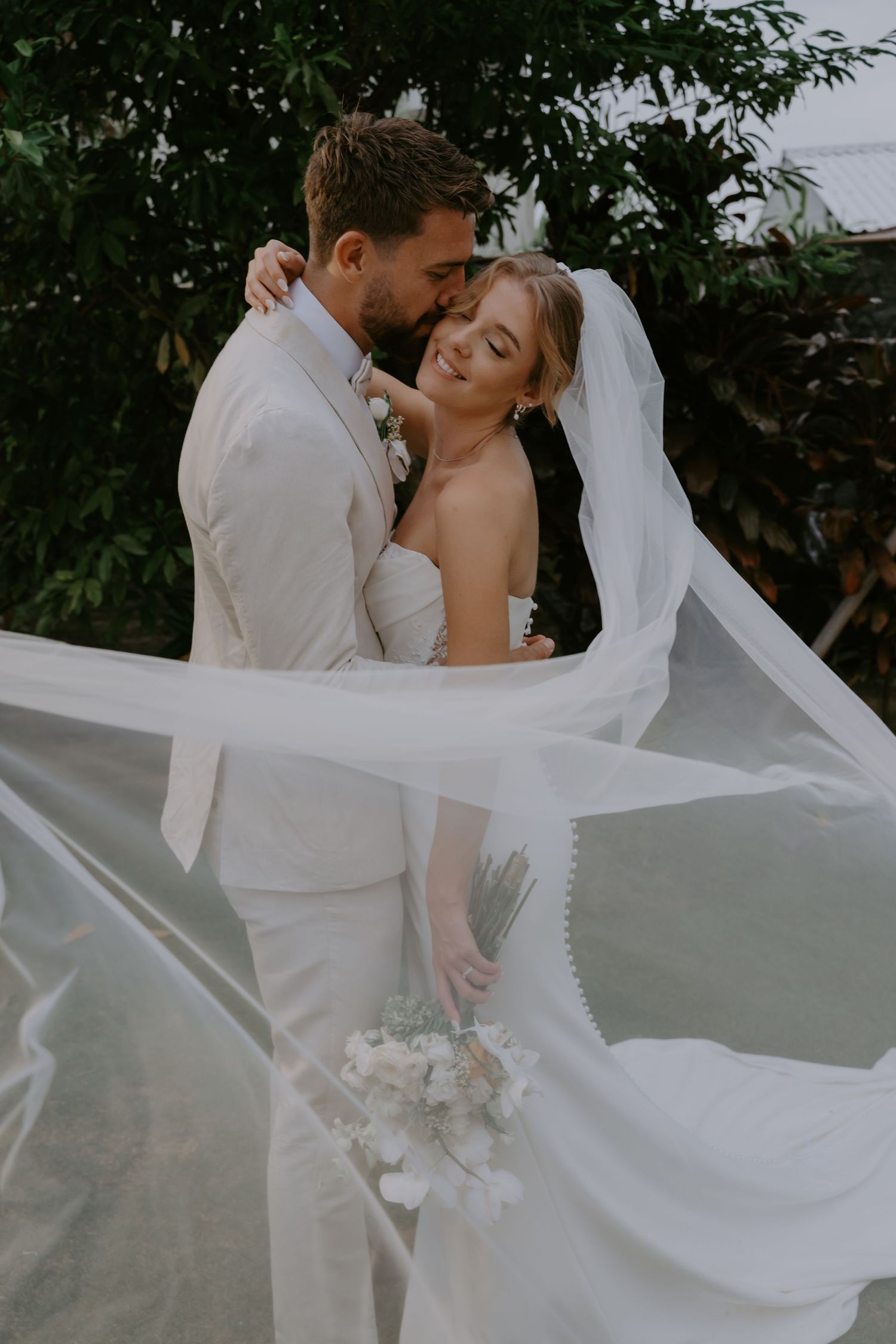 Bride and groom embrace outdoors, veil flowing in the wind, bride holding white bouquet in wedding attire.