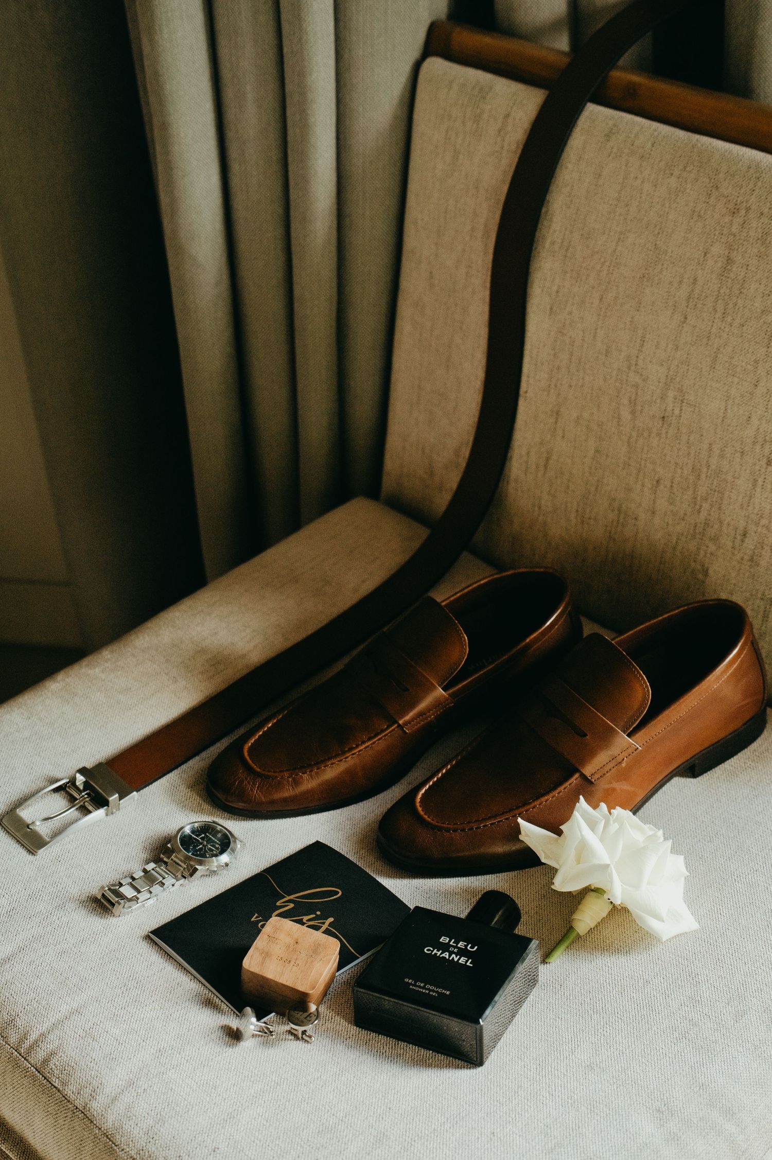 Brown leather penny loafers, Bleu de Chanel cologne, watch, cufflinks, and white rose on a beige chair.