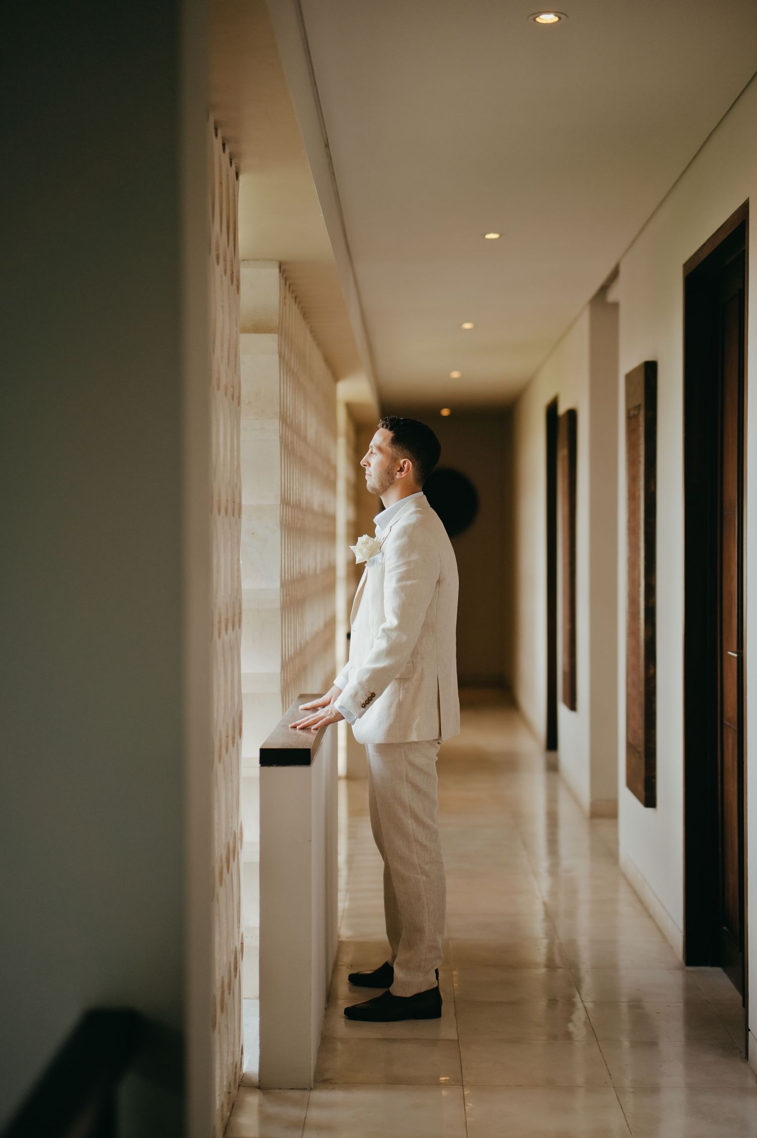 Groom in white linen suit stands thoughtfully in a modern hotel corridor before the ceremony.