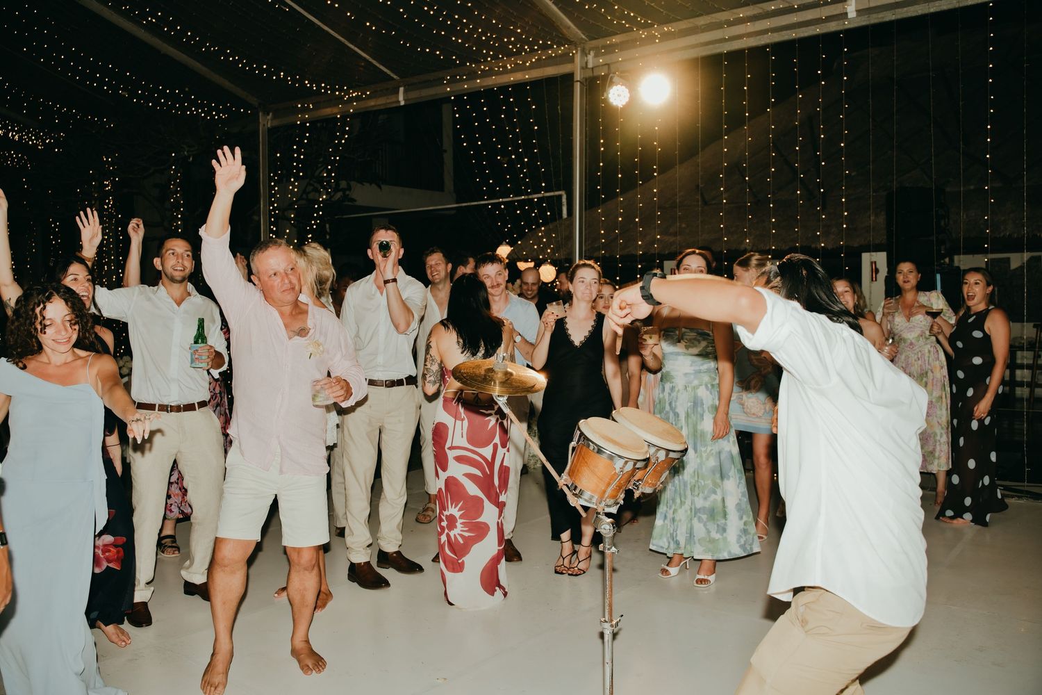 Energetic wedding reception dance floor with guests dancing and a live drummer performing under string lights.