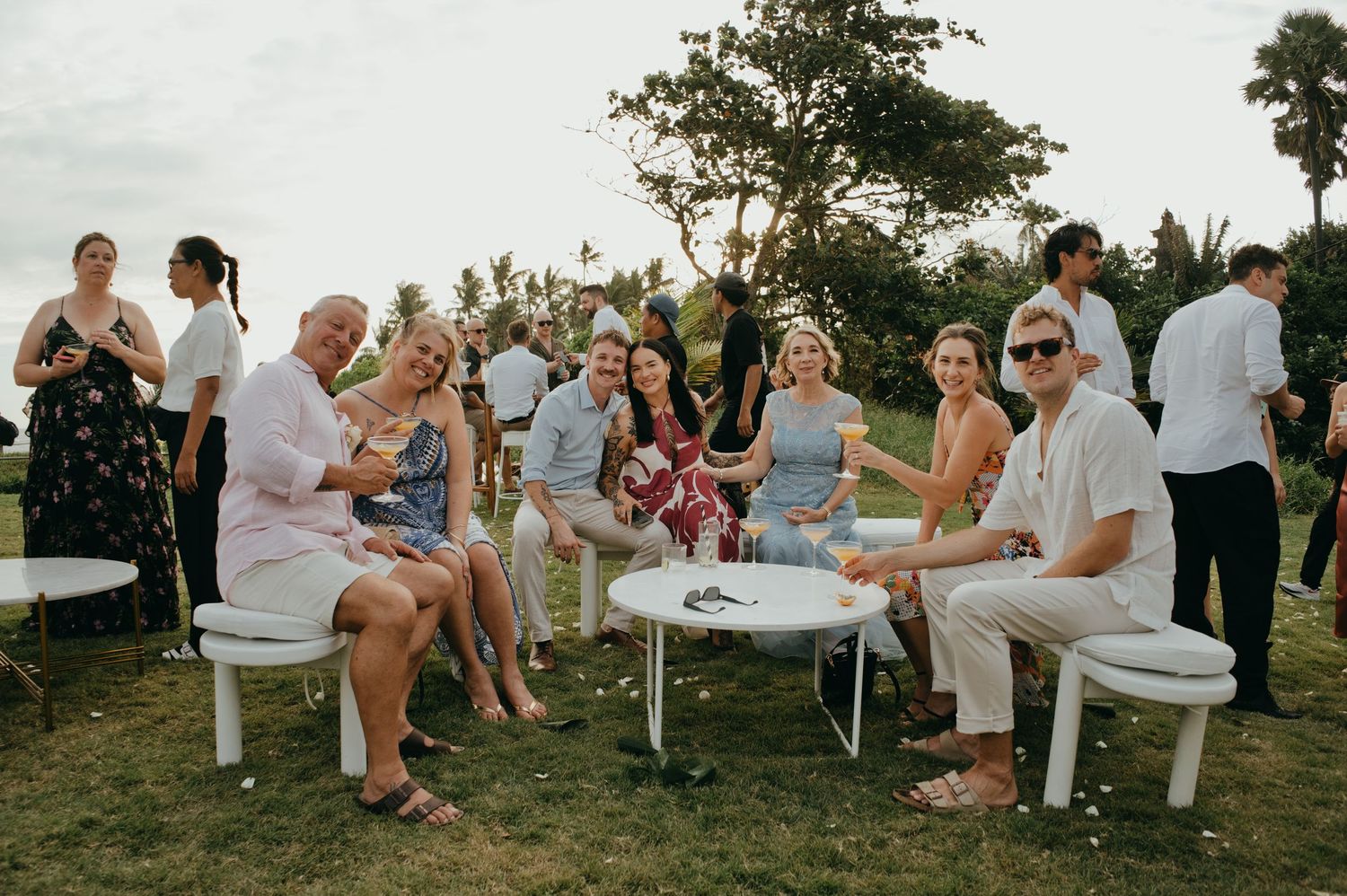 Wedding guests relax and socialize on white outdoor furniture at a garden cocktail hour.