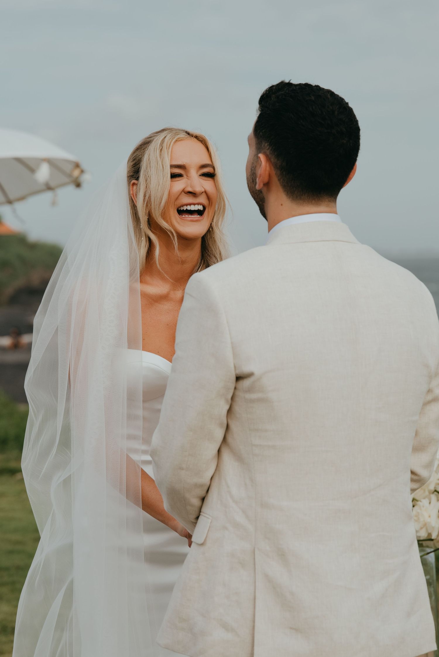 Laughing bride in white strapless wedding dress and veil exchanging vows with groom outdoors.
