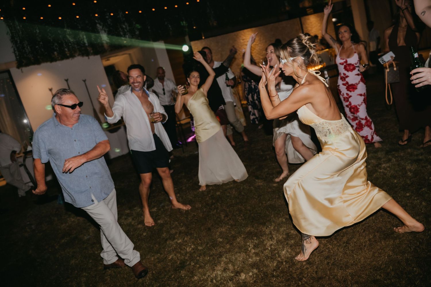 Bride and wedding guests dance energetically on the lawn at an outdoor evening wedding reception celebration.