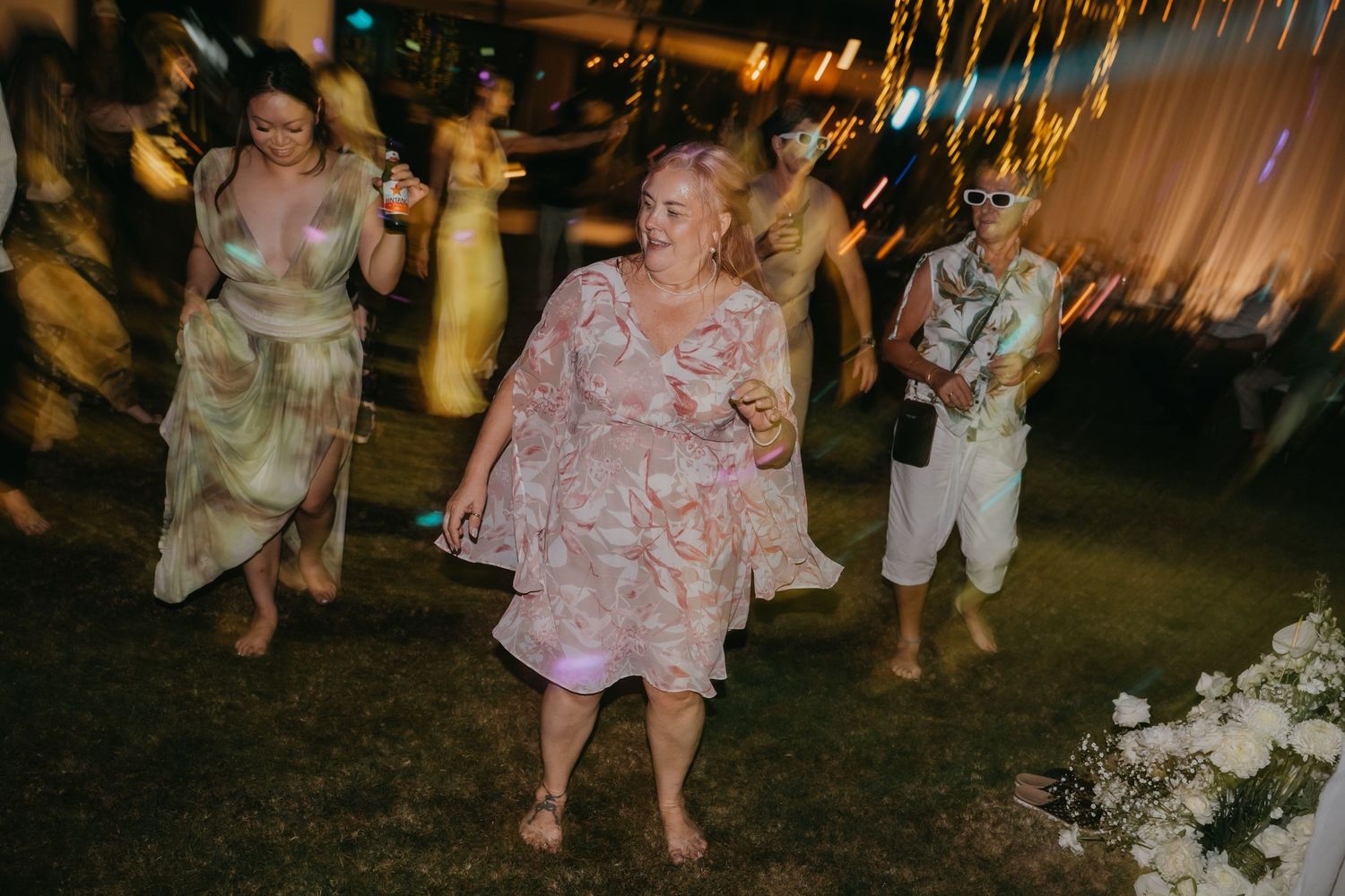 Guests dance joyfully on grass at outdoor evening wedding reception under warm festive lighting.