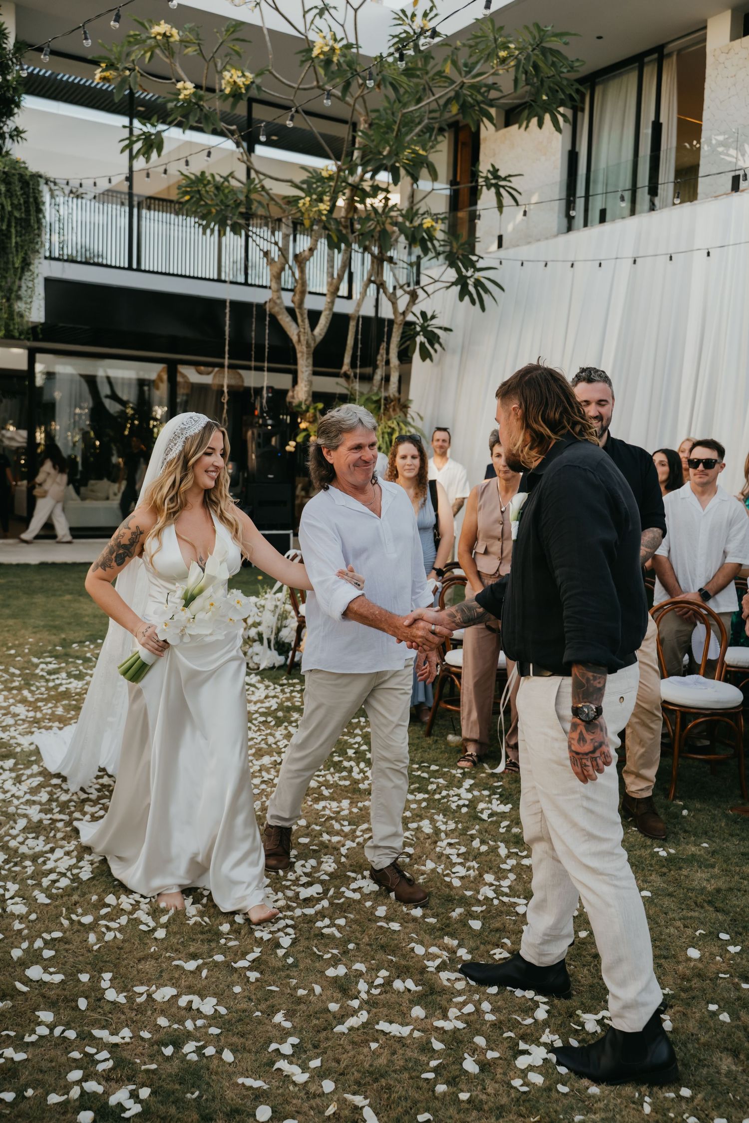 Bride walks down aisle escorted by father toward groom on flower petal-lined outdoor wedding ceremony.